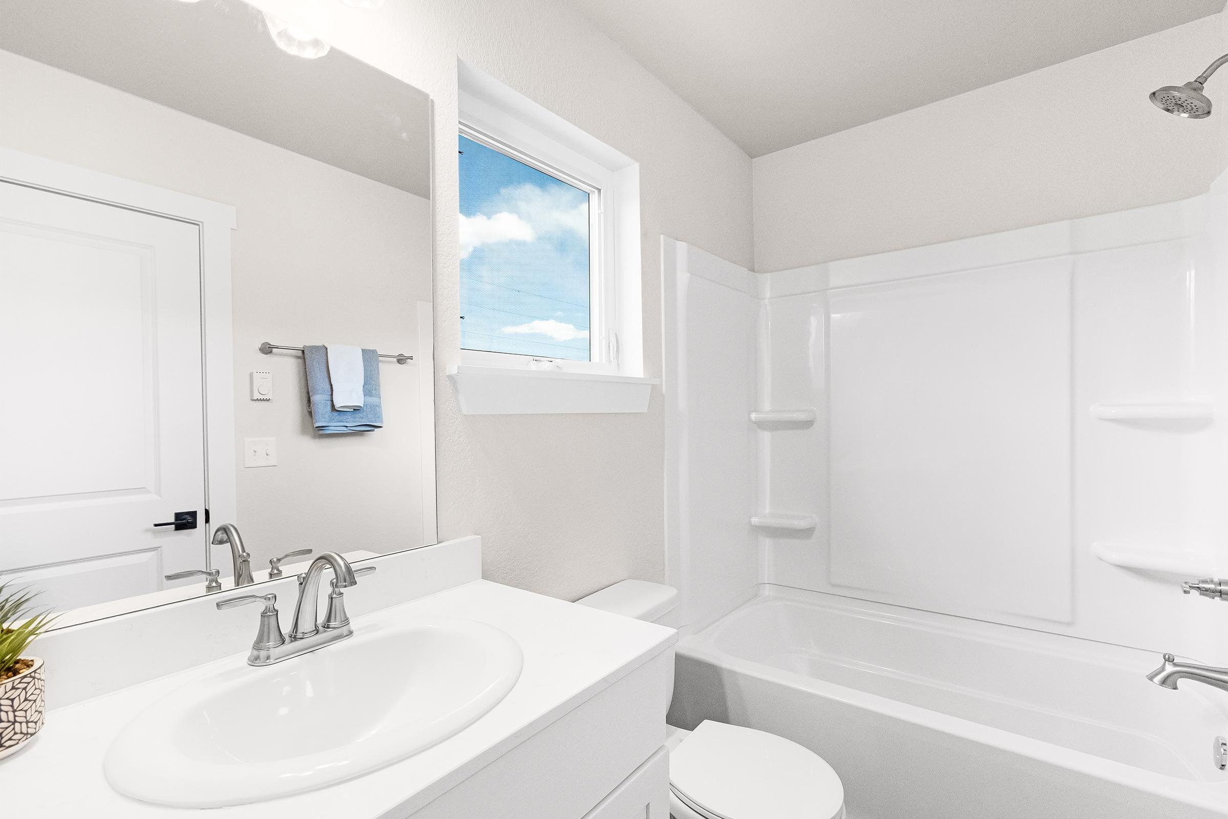 Bright and modern bathroom featuring a white bathtub and shower, a white vanity with a sink, and a large mirror. A window with a view of blue sky and clouds provides natural light, while light-colored walls create an airy feel. Light blue towels hang on a hook nearby.