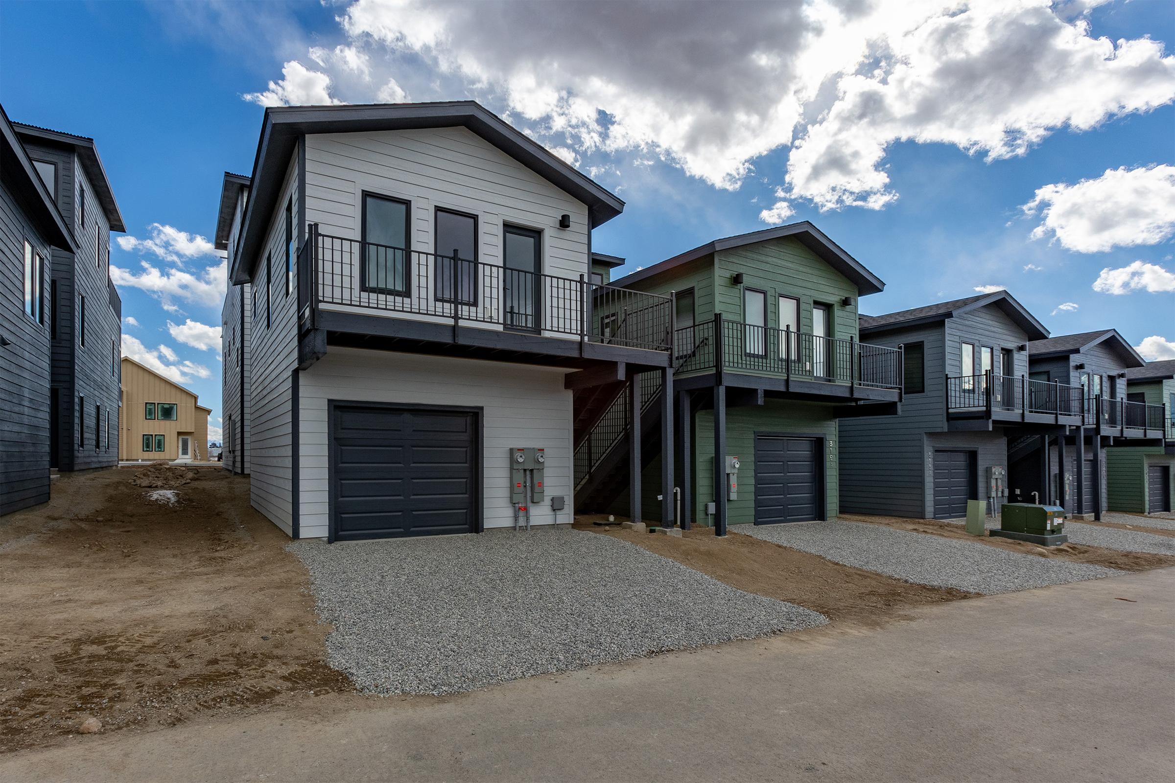 A row of modern two-story houses featuring a mix of dark and light siding. Each house has a garage, a small gravel driveway, and balconies. The sky is partly cloudy with blue and white clouds, and the surrounding area is still under development with dry ground visible.