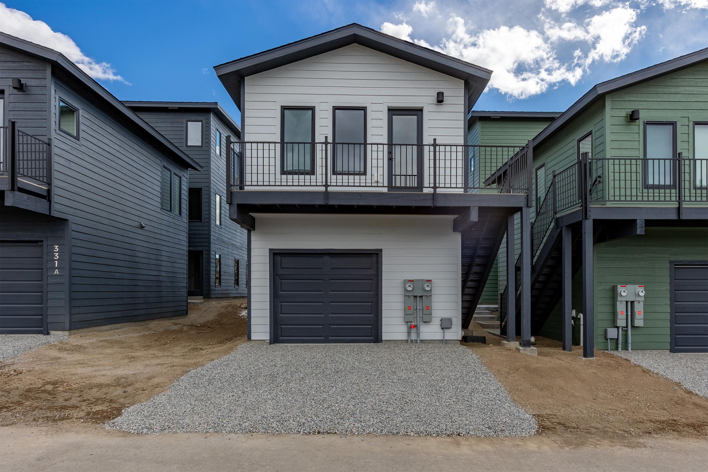 A modern-style house with a gray garage door, located between two darker-colored houses. The home features a white exterior, multiple windows, and a small balcony. A gravel driveway leads to the front, and utility meters are visible on the side. The sky is partly cloudy, adding a serene atmosphere.