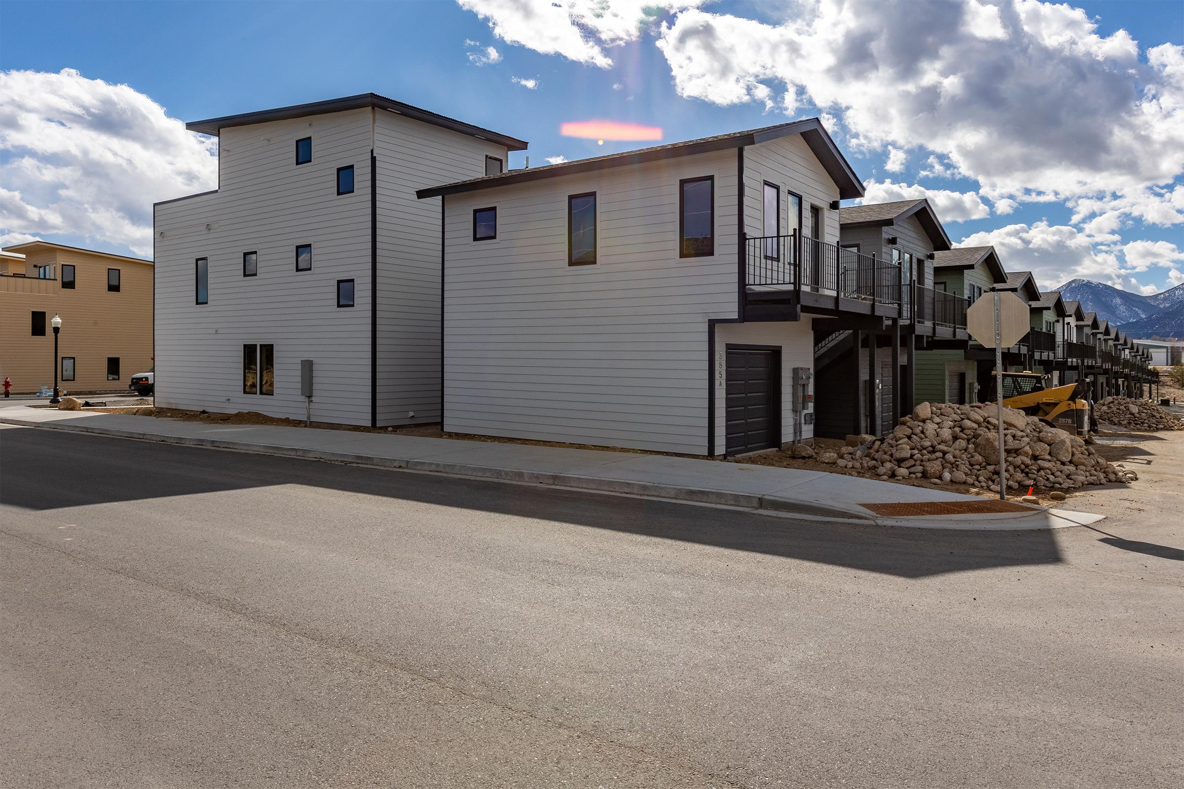 A modern residential neighborhood featuring two contemporary multi-story buildings with a mix of light and dark siding. The scene shows a newly developed area with unfinished construction, sidewalk, and some landscaping materials, under a partly cloudy sky with mountains in the background.
