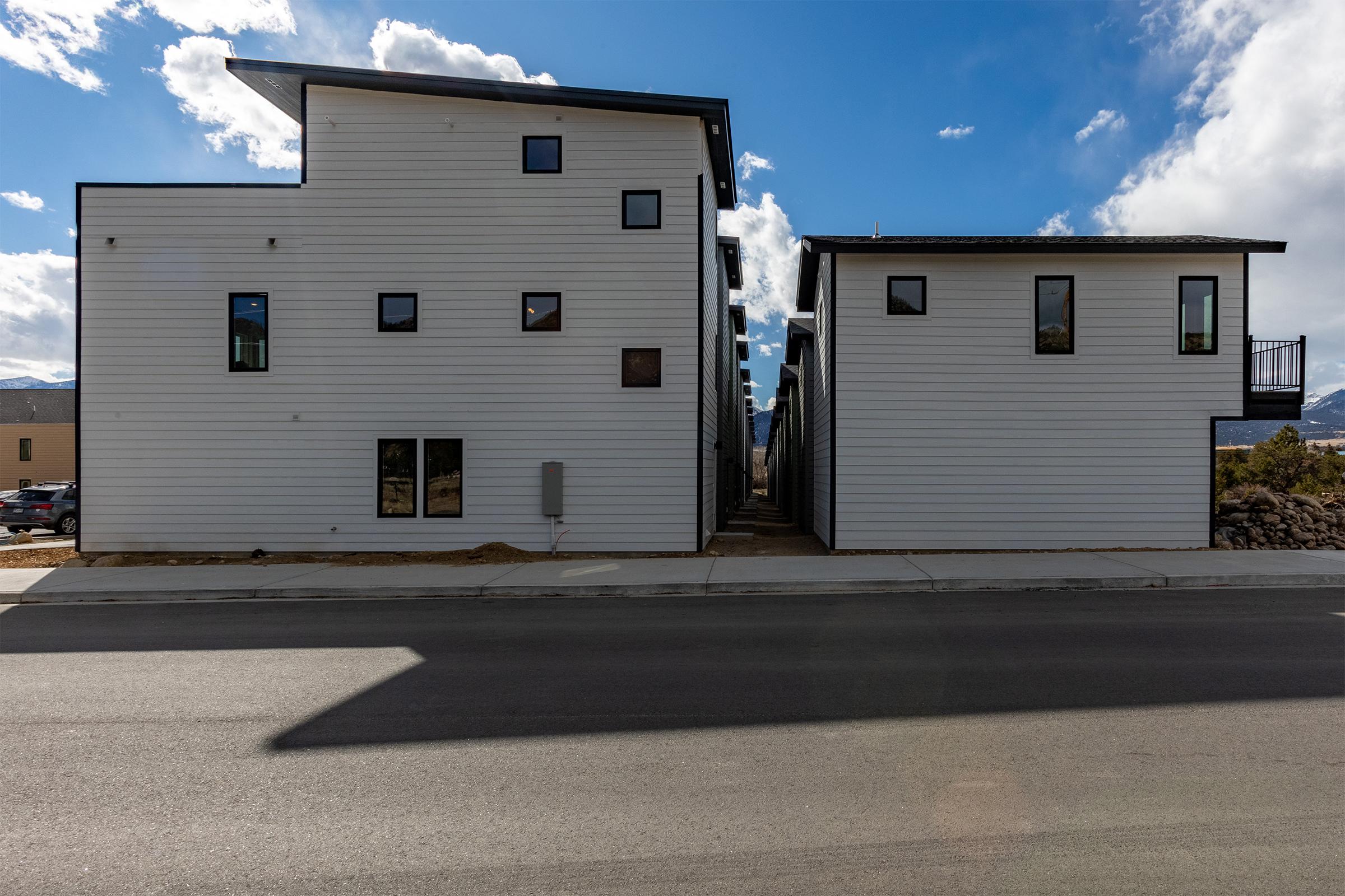 Two modern, two-story buildings with a white exterior and multiple windows. The buildings are separated by a narrow pathway, and the scene is set against a blue sky with scattered clouds. A paved road runs in front, and there are some vehicles parked nearby.