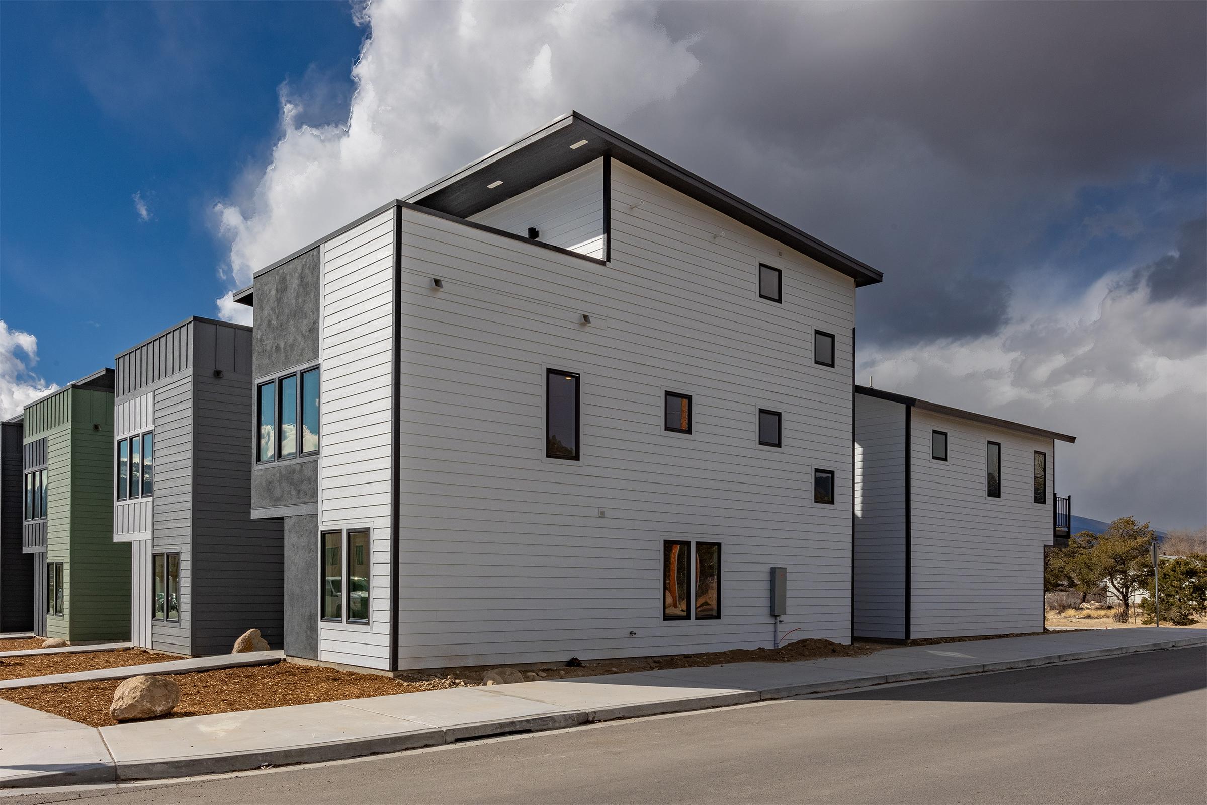 Modern two-story residential building with a mix of white and gray siding, large windows, and a contemporary design. The structure features angular lines and a minimalist aesthetic, set against a backdrop of partly cloudy skies. A paved sidewalk runs alongside the building in a suburban setting.