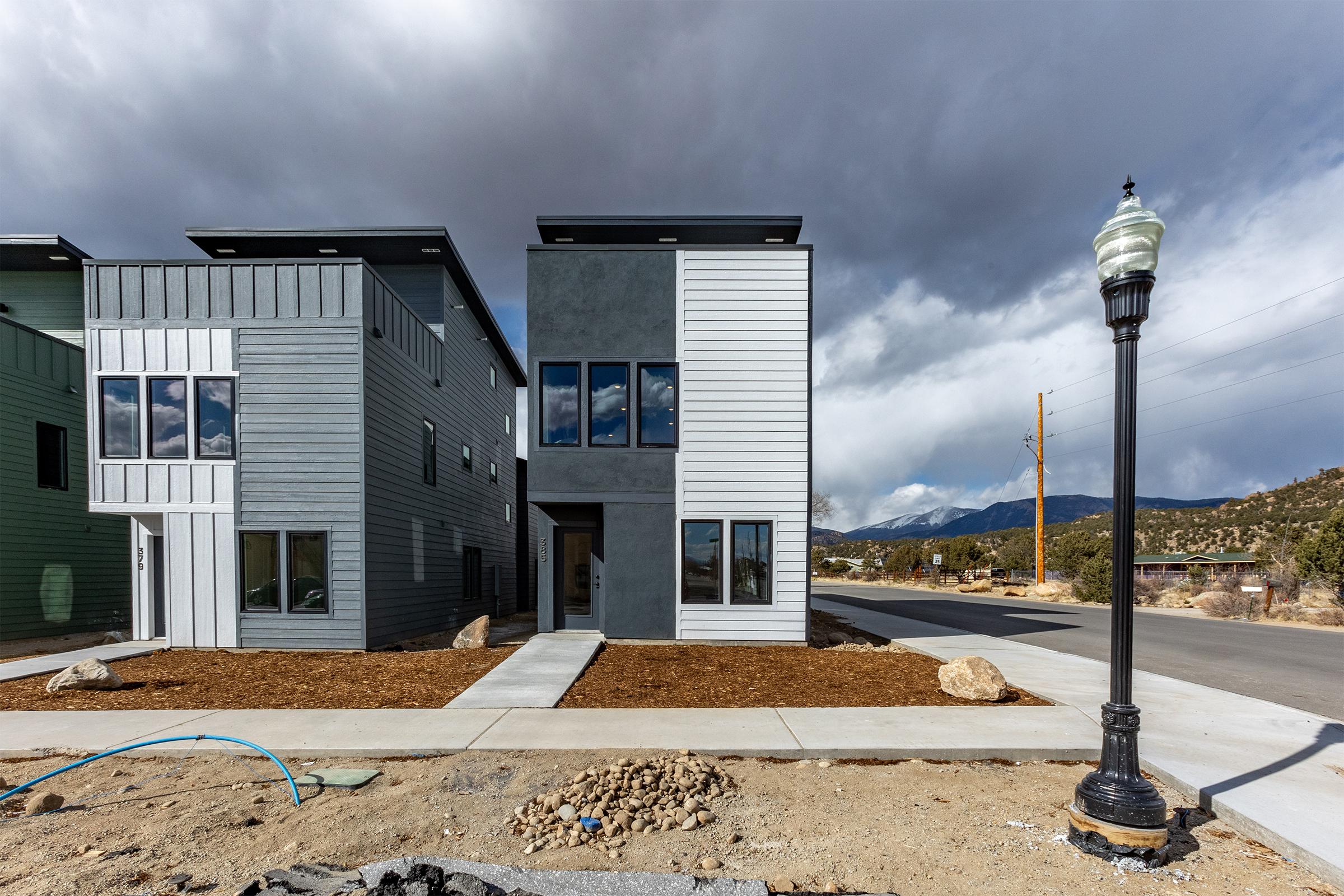 A row of modern, two-story houses with varied exterior finishes, including light gray and dark gray. The buildings feature large windows and clean lines. A lamp post stands on the sidewalk, while the surrounding area is under development with rocks and dirt visible. Mountains are visible in the background under a cloudy sky.