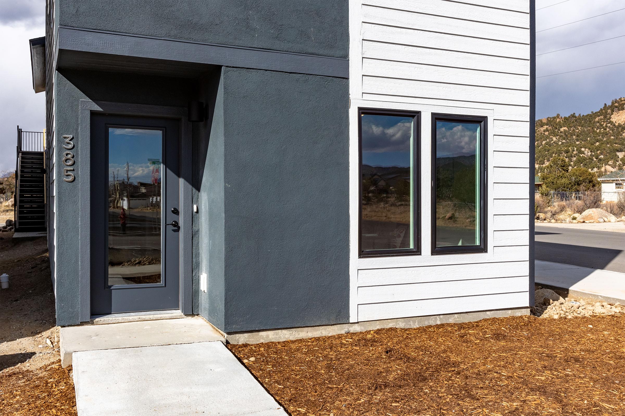 Modern two-story building exterior featuring a corner entrance with a grey door. The facade is divided between dark grey and light vertical siding. There are large windows on both sides of the entrance, and the landscaping includes a gravel pathway and mulch around the base of the building, with a mountainous landscape in the background.