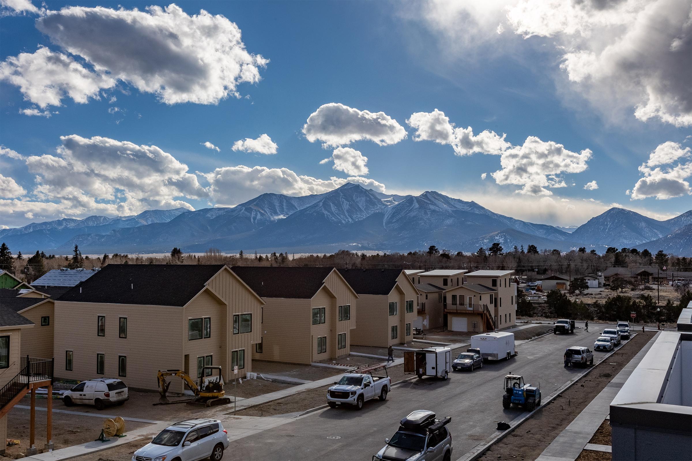 A scenic view of a mountainous landscape with dramatic clouds. In the foreground, there are residential buildings under construction alongside a street with parked vehicles. The mountains rise majestically in the background, capturing the beauty of the natural surroundings.