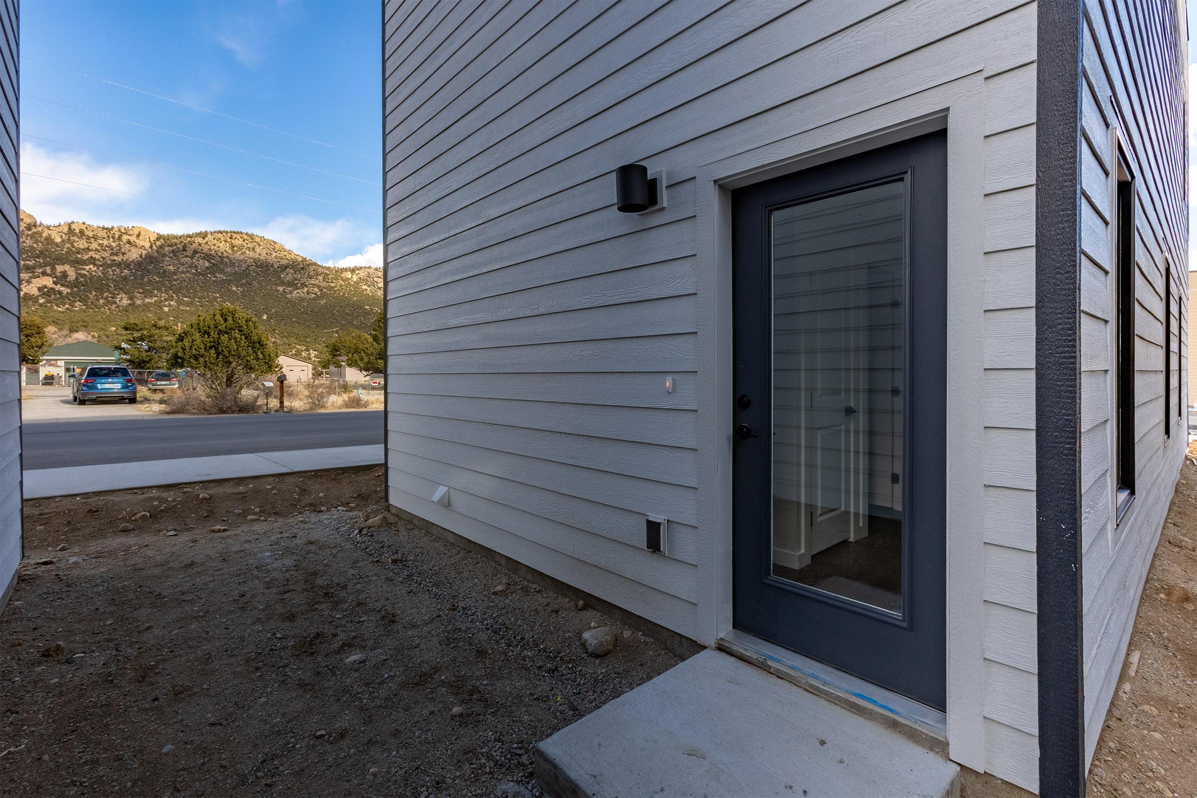 A corner view of a modern building featuring a gray door with a glass panel. The structure has light-colored horizontal siding and is situated in a rural area with mountains in the background. A gravel path leads to the entrance, and trees and vehicles are visible in the distance.