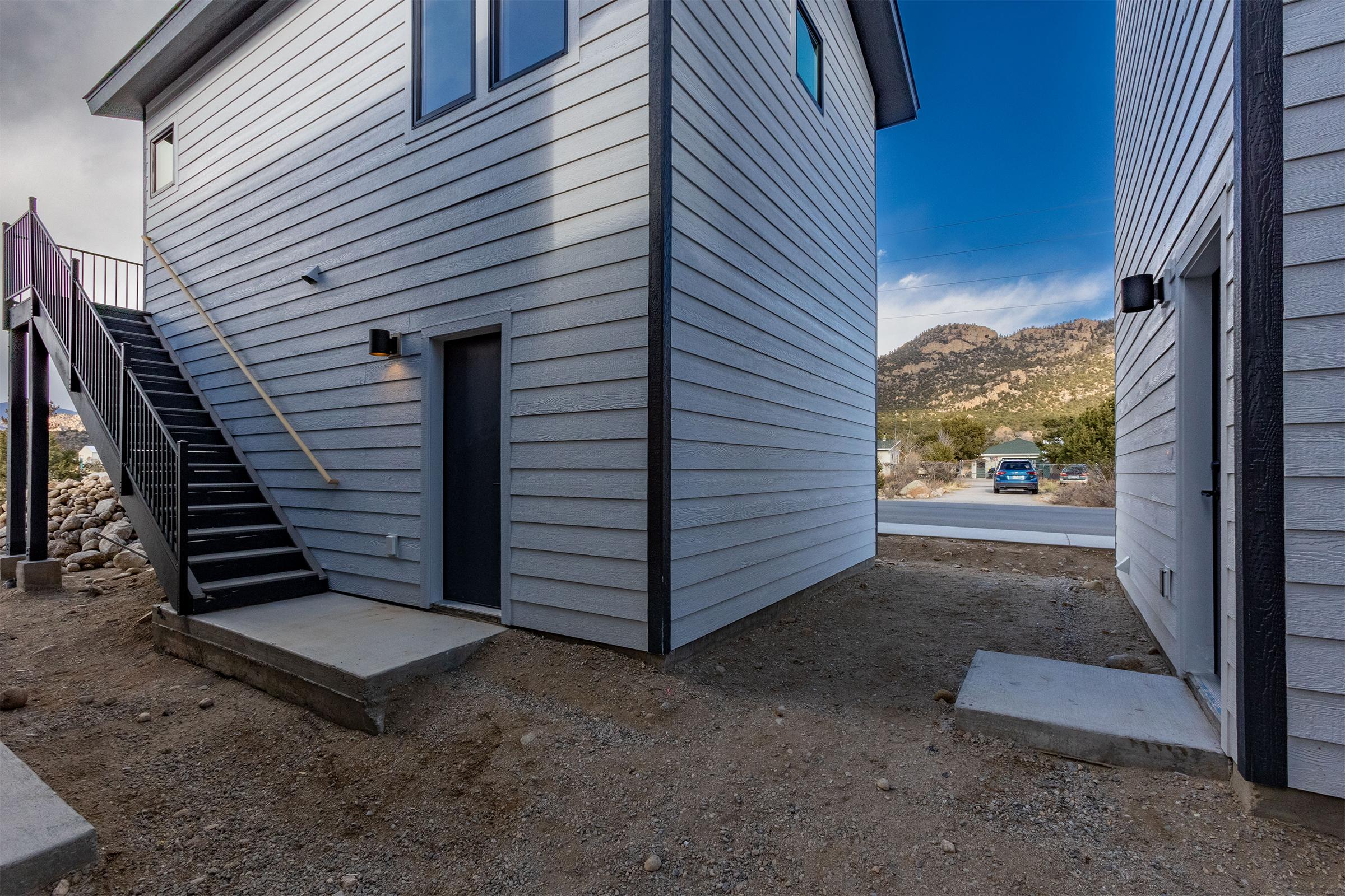 Two modern, gray, two-story buildings are positioned closely together, with a set of external stairs leading to the upper level of one. The ground is rocky and bare, and there are utility poles in the background. A blue vehicle is visible in the distance along a road surrounded by mountains.
