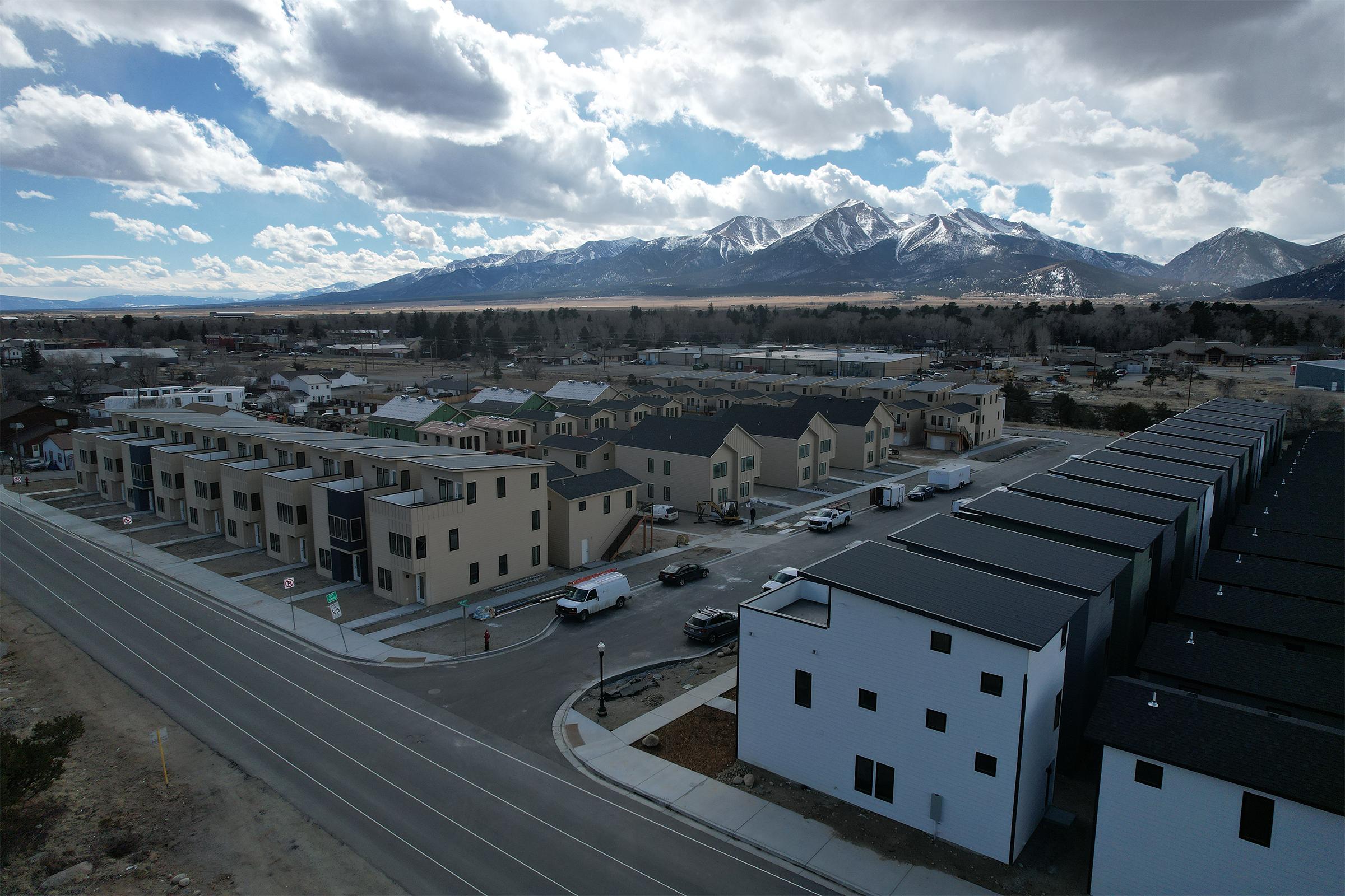 Aerial view of a residential area featuring modern multi-story buildings and townhouses, with a clear road in the foreground. In the background, there are snow-capped mountains under a partly cloudy sky, showcasing a mix of urban development and natural landscape.