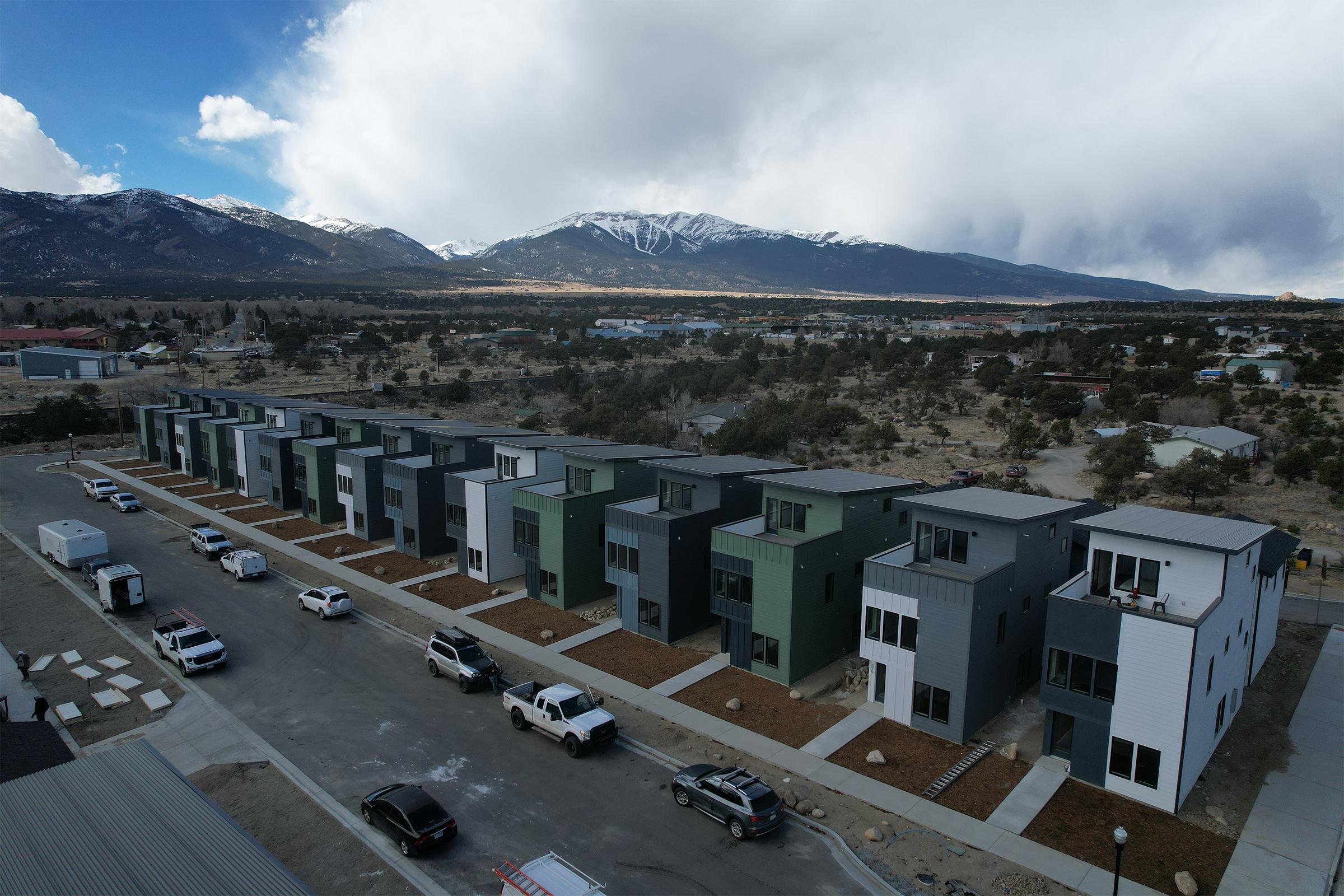 Aerial view of a row of modern, multi-colored houses along a paved road. Snow-capped mountains loom in the background under a cloudy sky. Several parked vehicles are visible in the foreground, and the area features sparse vegetation typical of a semi-rural landscape.