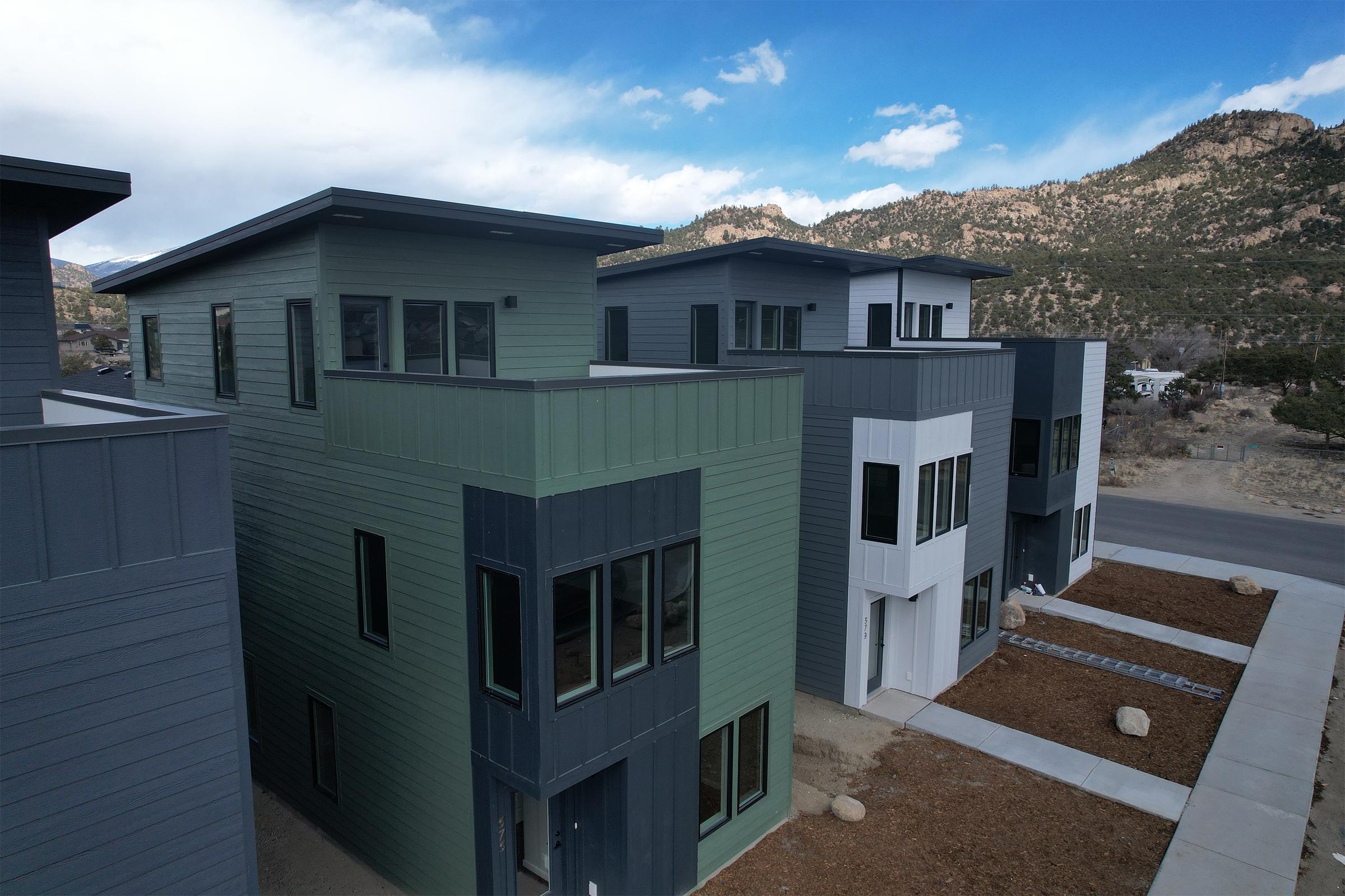 A row of modern, multicolored houses with a mix of gray, green, and white exteriors. The buildings feature large windows and flat roofs, set against a backdrop of rocky hills and a blue sky with scattered clouds. A paved walkway and landscaped area with gravel and boulders are in the foreground.