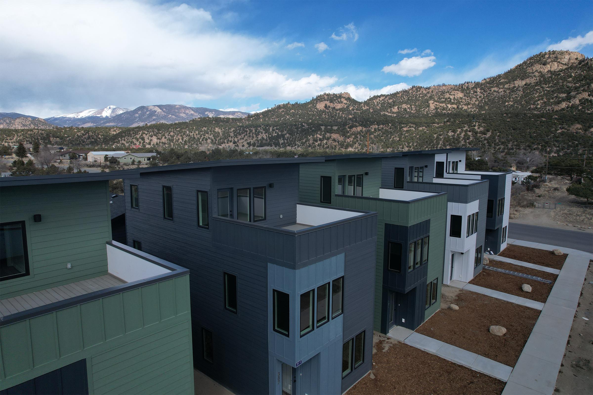 Modern residential buildings with a mix of dark and light façades, situated in a scenic mountainous area. The sky features some clouds, and the landscape includes trees and hills, creating a serene environment. The foreground shows a pathway leading to the buildings, with some bare landscaping.