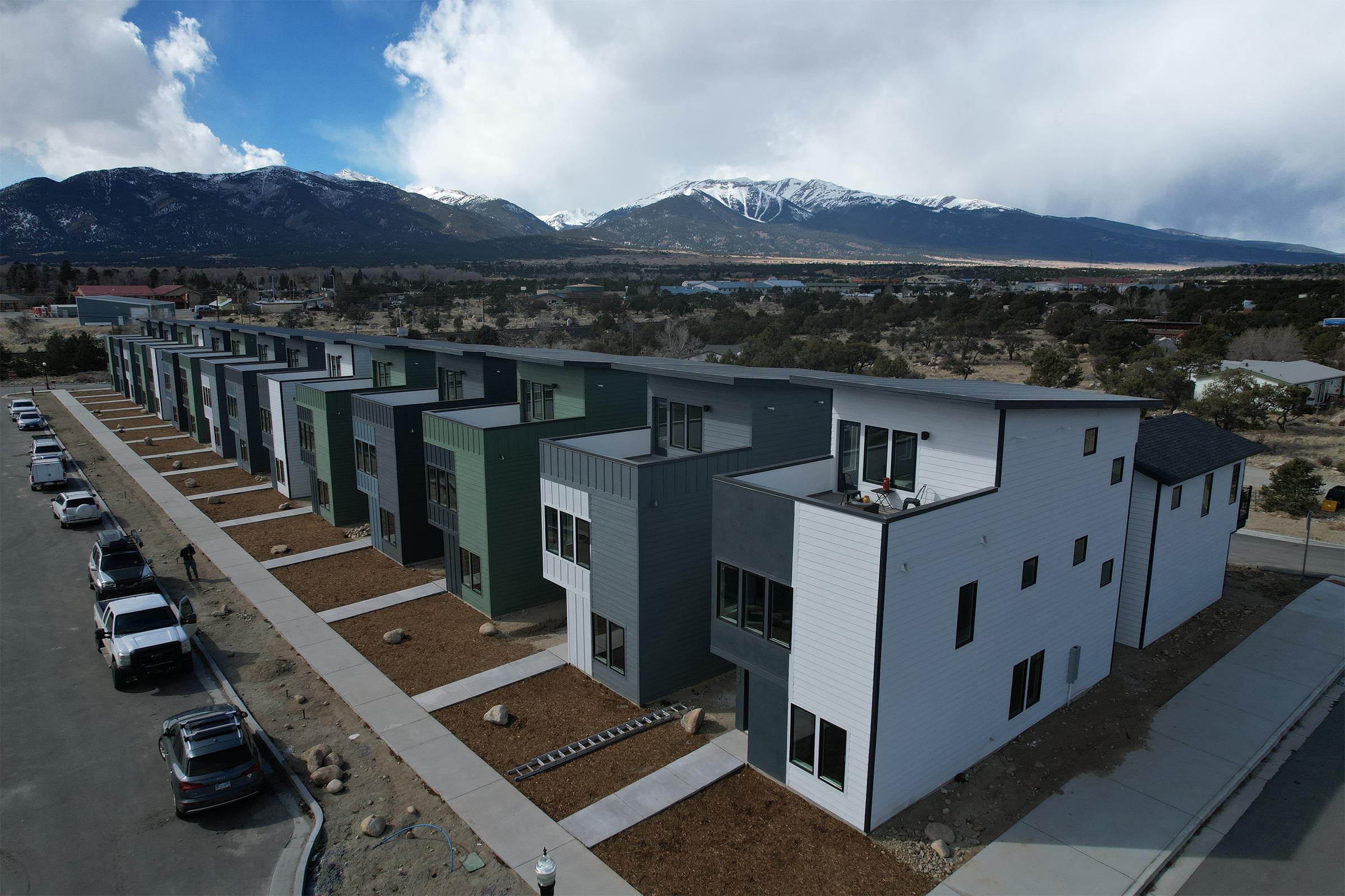 A row of modern, multi-story homes with a mix of green and gray exteriors. The buildings are situated in a rural area with mountains in the background, under a partly cloudy sky. Asphalt parking spaces are visible in front of the homes, along with landscaped patches of gravel and rocks.