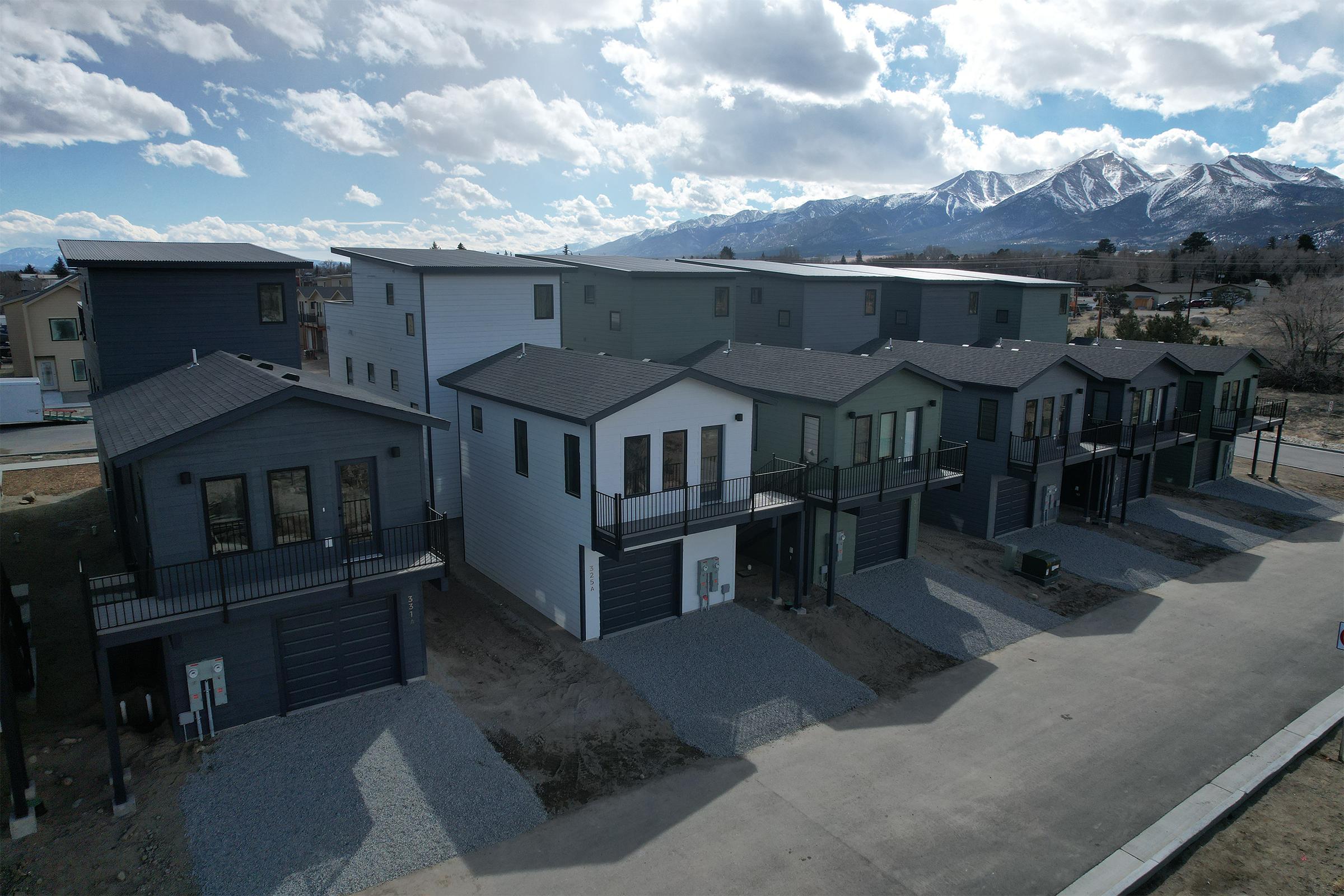 A row of modern, multi-story houses with various styles and colors, featuring large windows and balconies. The homes are set against a backdrop of mountains under a partly cloudy sky. Gravel driveways are visible in front of the houses. The landscape appears newly developed.
