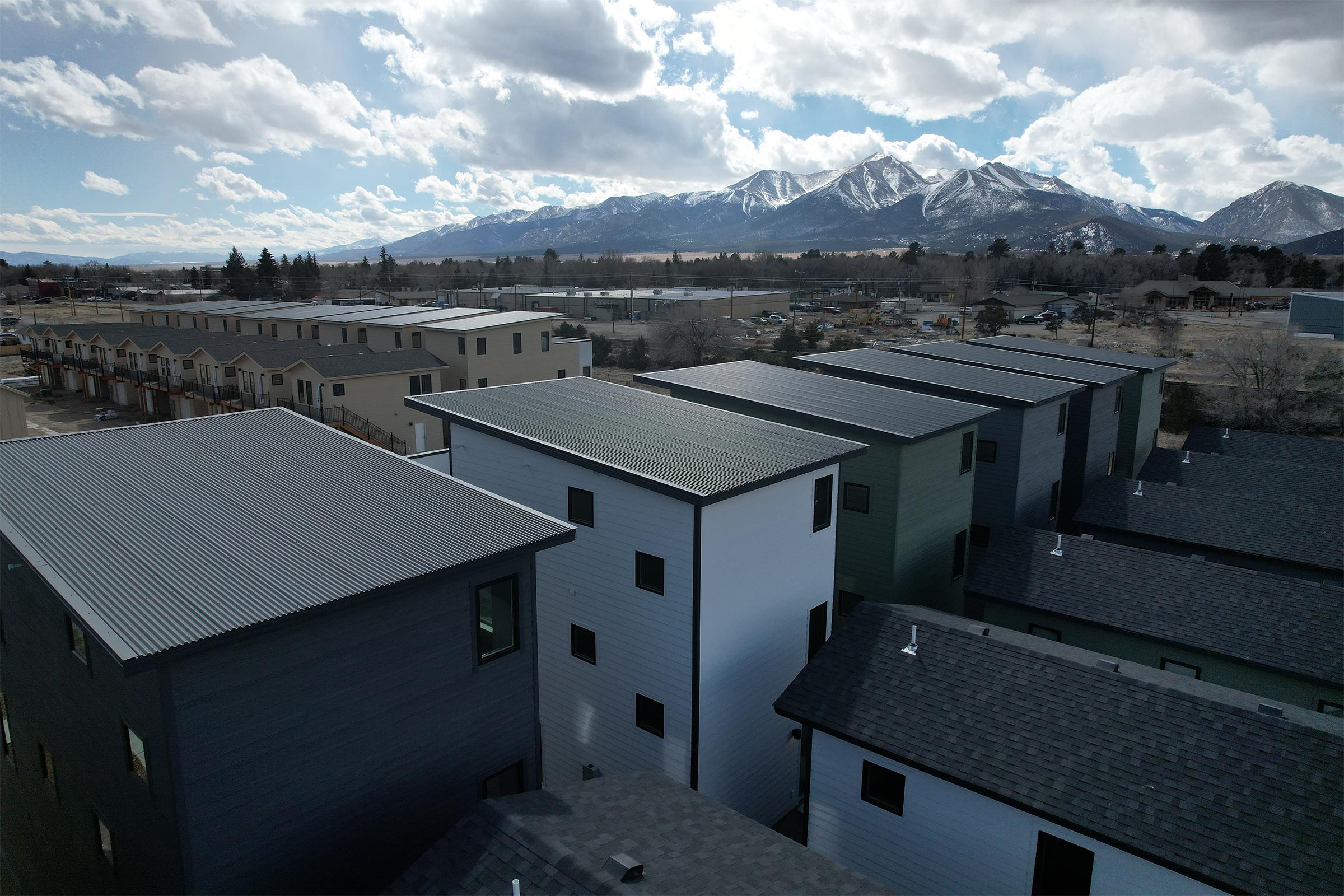 Aerial view of modern homes featuring flat roofs, with solar panels visible. The buildings are nestled in an urban area, surrounded by a mountainous landscape under a partly cloudy sky. The scene highlights a blend of contemporary architecture and natural beauty.