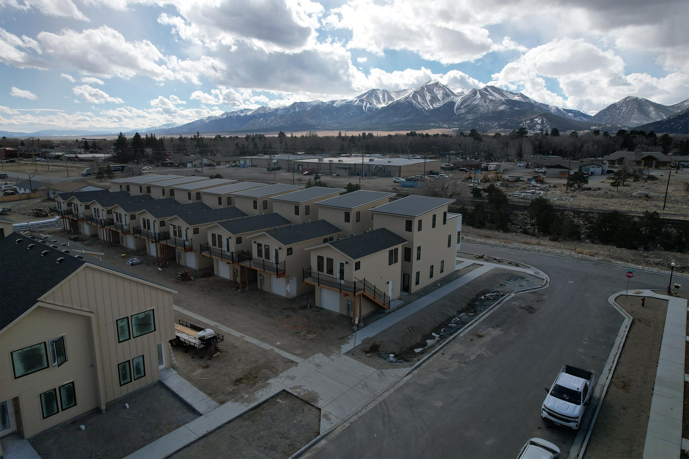 Aerial view of a residential area featuring several new, beige-colored townhouses with multiple units. In the background, majestic mountains are visible under a partly cloudy sky. The foreground shows a recently paved road and some construction materials, indicating ongoing development in the area.