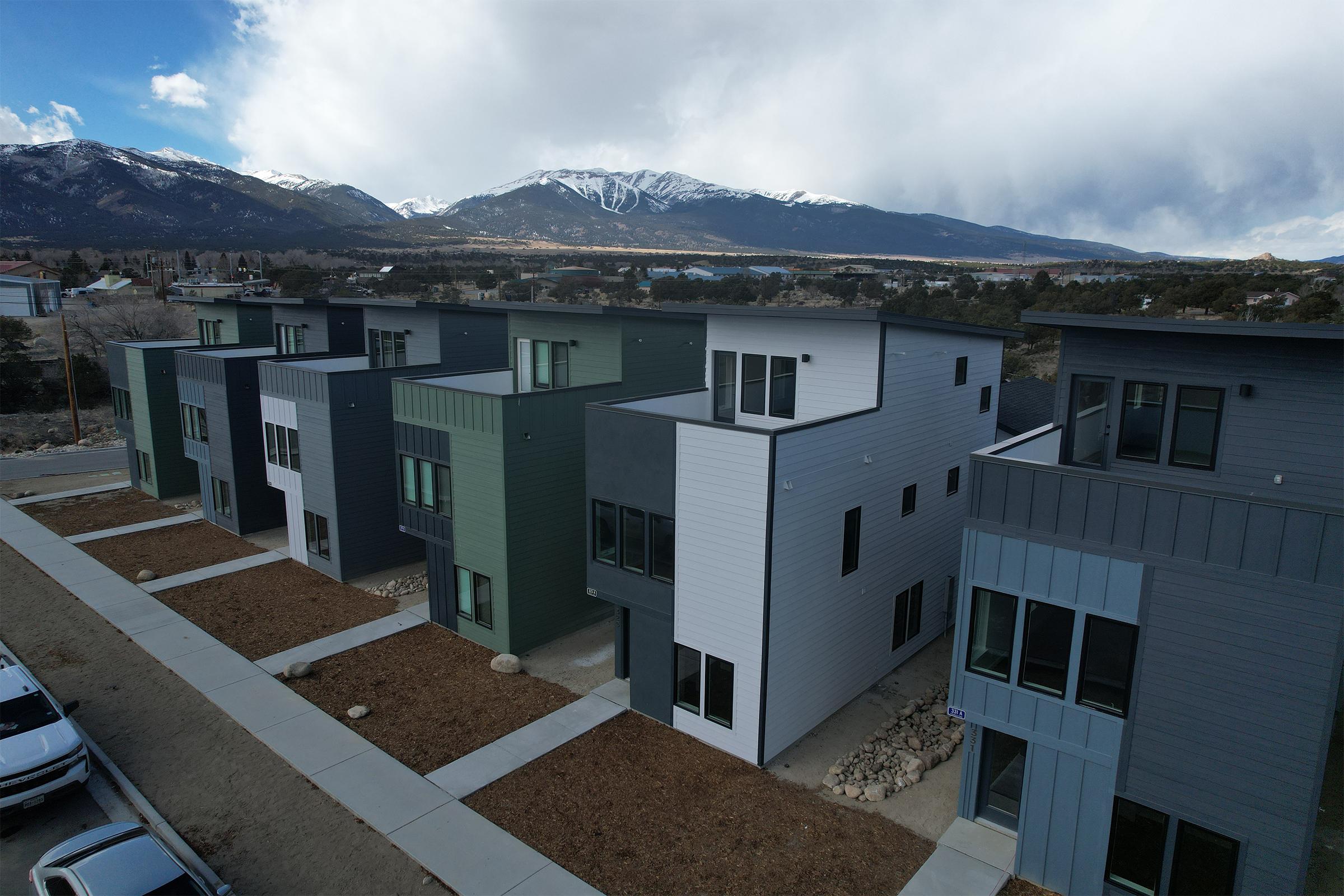 Aerial view of modern, multi-story residential buildings with varying colors (green, gray, white) alongside a gravel pathway. Snow-capped mountains are visible in the background under a partly cloudy sky, with a lake in the distance. The scene conveys a blend of nature and contemporary architecture.