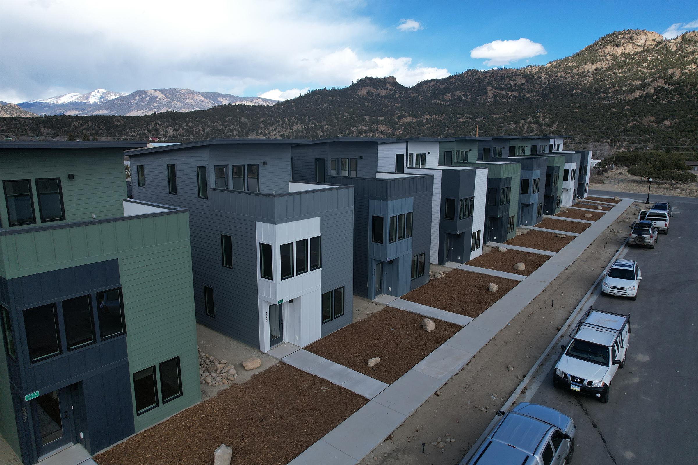 Aerial view of a modern residential complex featuring multiple gray and blue townhouses. The buildings are arranged in a row with landscaped areas and a gravel pathway in front. Rocky mountains and a partly cloudy sky can be seen in the background. Several parked vehicles line the street beside the complex.