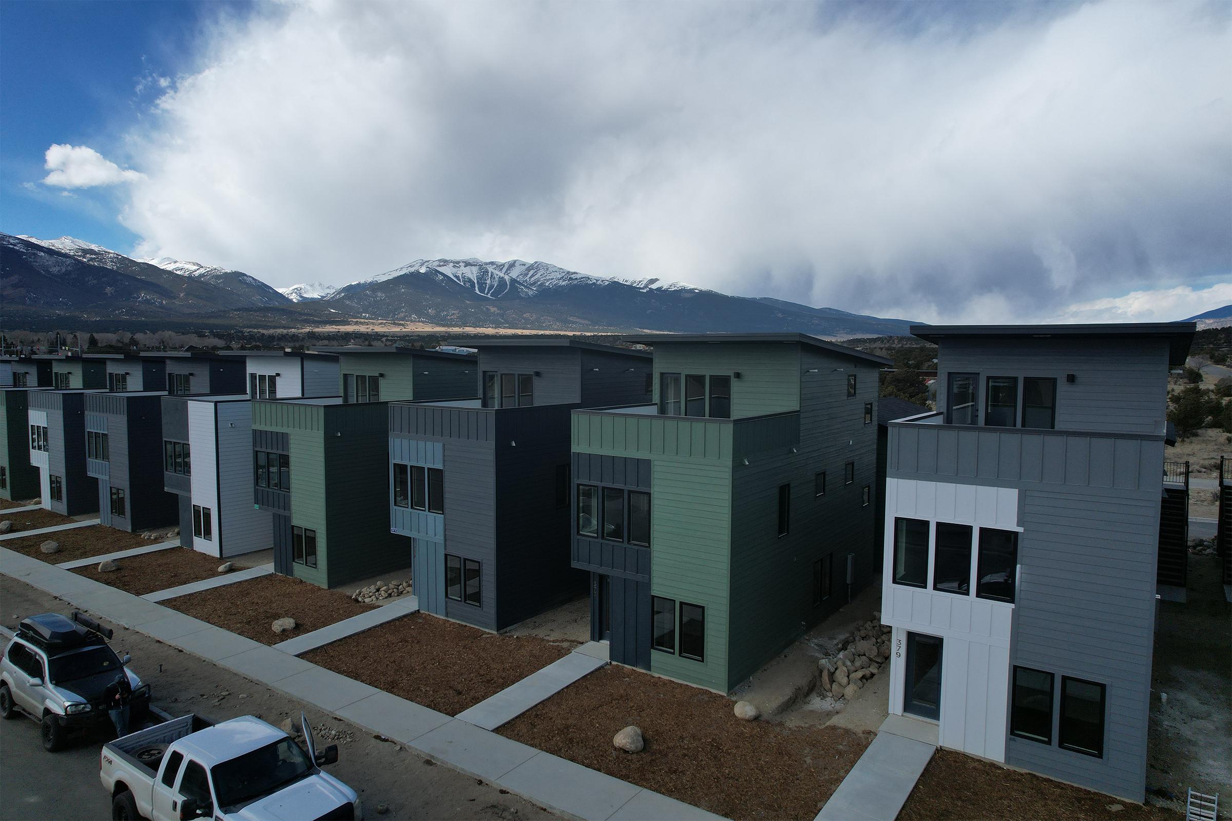 Row of modern, multi-story homes in various shades of gray and green, set against a backdrop of mountains and a partly cloudy sky. The neighborhood features landscaped paths and parked vehicles.