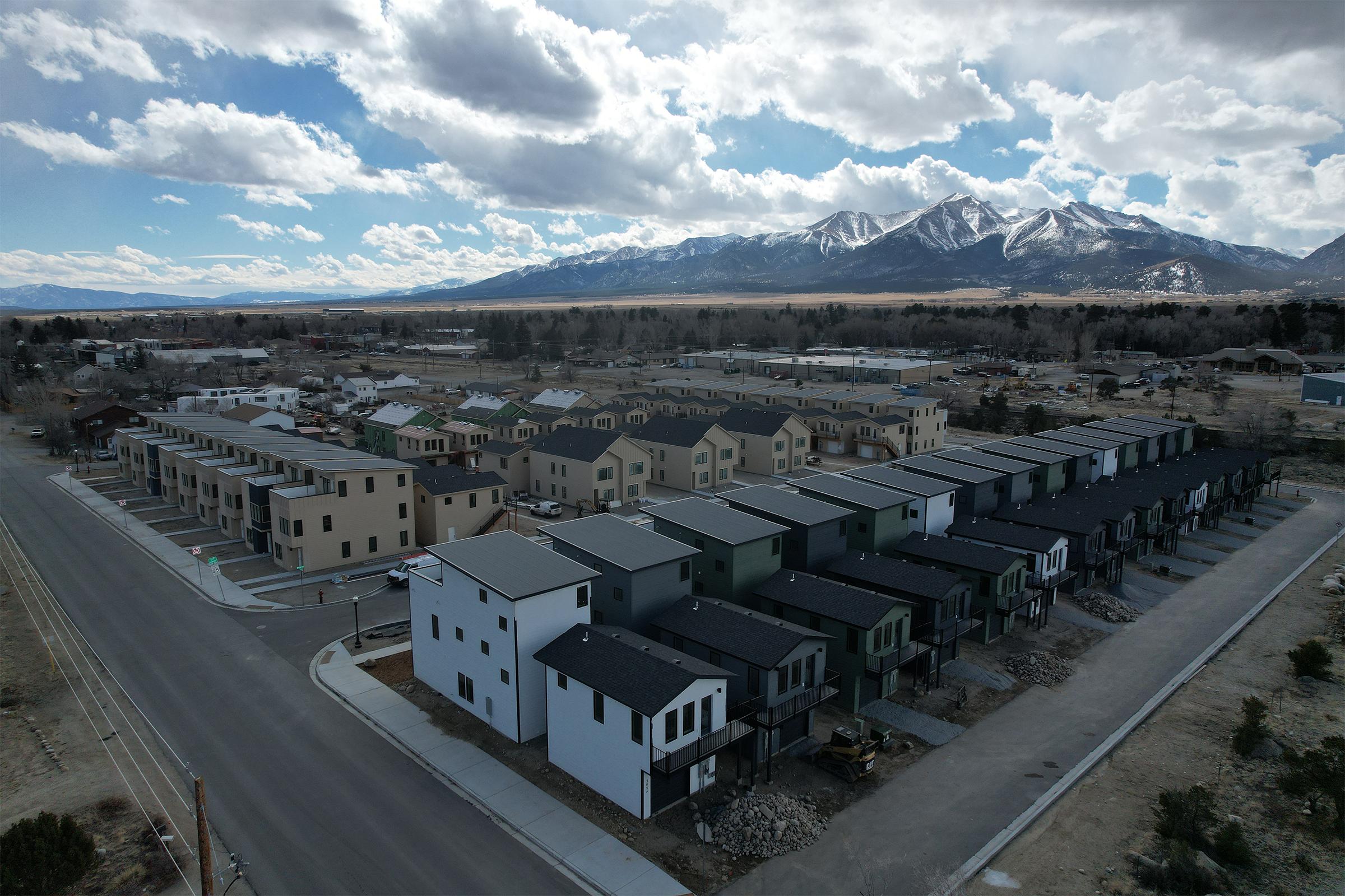 Aerial view of a residential neighborhood featuring modern multi-family homes with varying colors. In the background, there are snow-capped mountains and a wide open landscape under a partly cloudy sky. The streets are paved and lined with parked cars, showcasing a well-planned community layout.
