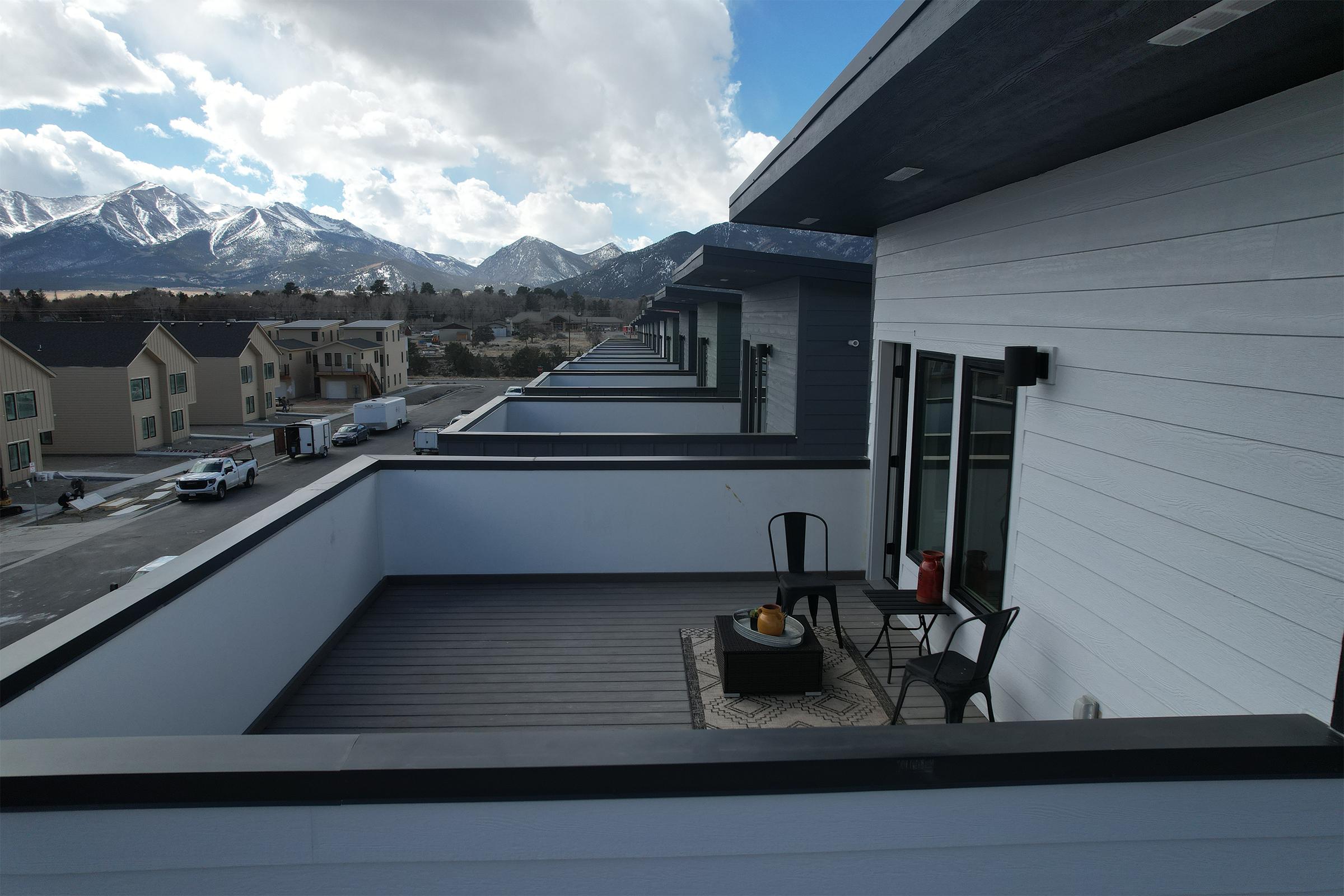 A view of modern residential buildings with balconies, overlooking a mountain range under a partly cloudy sky. The scene features a spacious balcony with outdoor furniture and a decorative rug, while additional homes line the street below.
