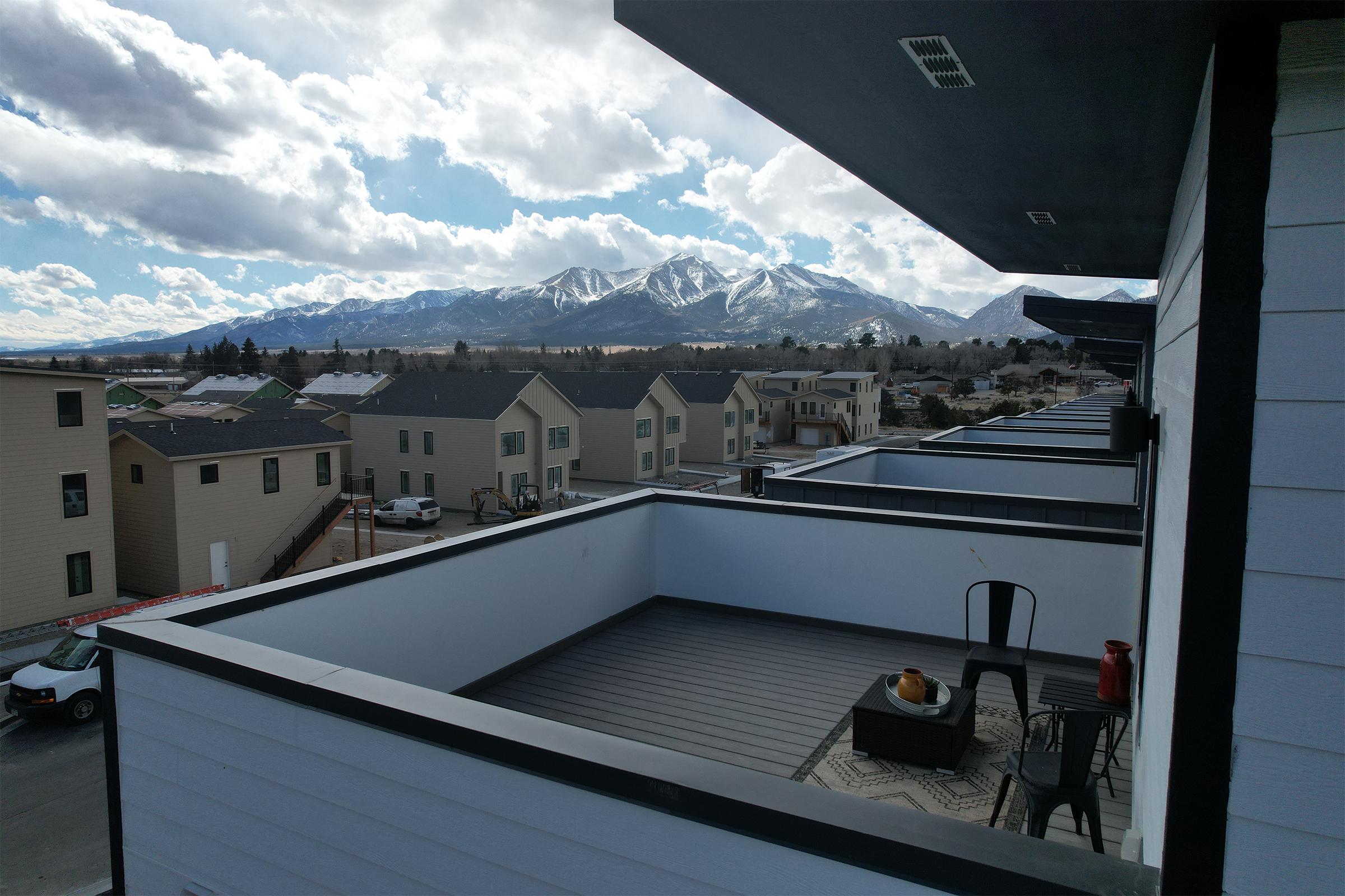 A view from a balcony overlooking a residential area with modern houses and a distant mountain range under a partly cloudy sky. The balcony features a small table and chair, suggesting a comfortable outdoor space.