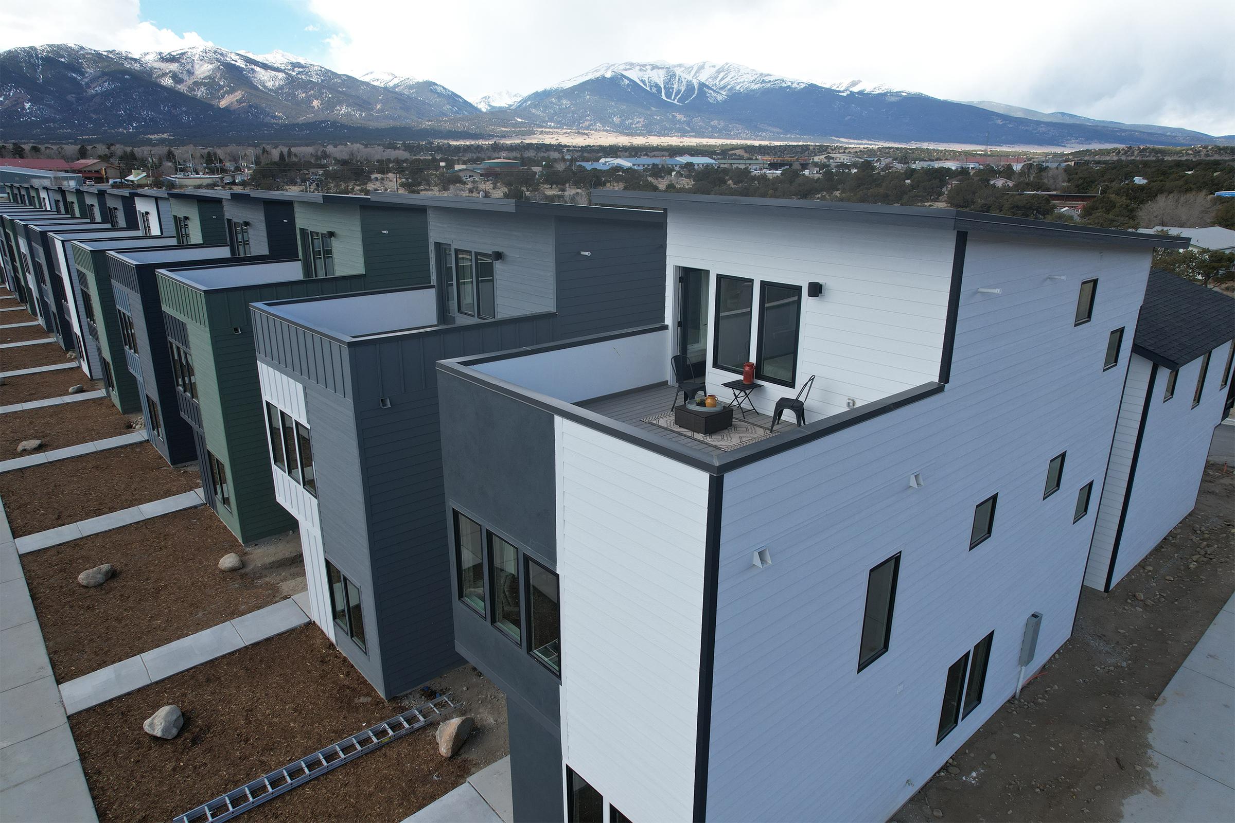 Aerial view of modern, multi-unit residential buildings with a mix of black, gray, and white siding. Each unit features large windows and rooftops. The background shows snow-capped mountains and a valley, with some landscaping and rocks visible in the foreground.