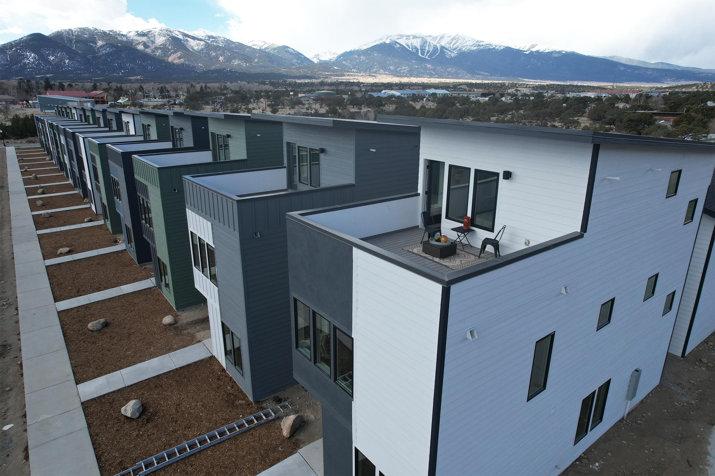Row of modern multi-story buildings in a residential area, featuring a mix of colors like green, gray, and white. The buildings are designed with balconies, and there’s a mountain range in the background under a partly cloudy sky. A construction ladder and landscaped area are also visible.