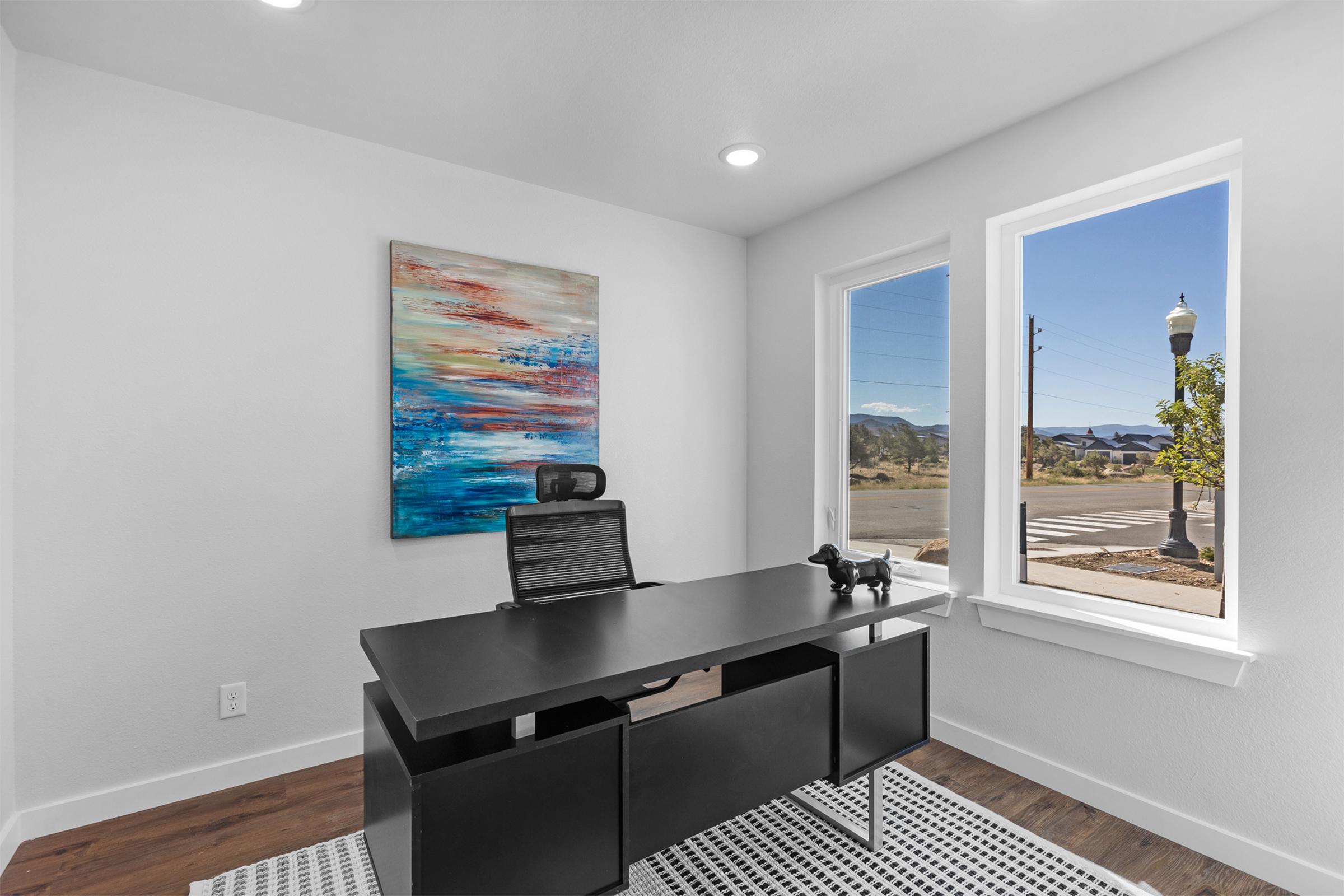A modern office setup featuring a sleek black desk with a chair, positioned near two large windows. One window offers a view of a street and mountains in the distance. A colorful abstract painting hangs on the wall, and a small black decorative object is on the desk. The floor is covered with a patterned rug.