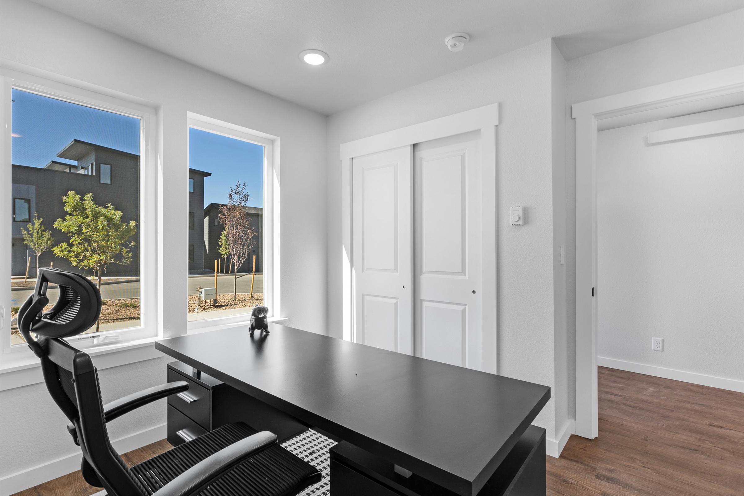 A modern office space featuring a sleek black desk and ergonomic chair. The room has large windows providing natural light, with views of trees and neighboring buildings outside. The walls are painted white, creating a bright and airy atmosphere. A pair of closed white doors is visible on the right.