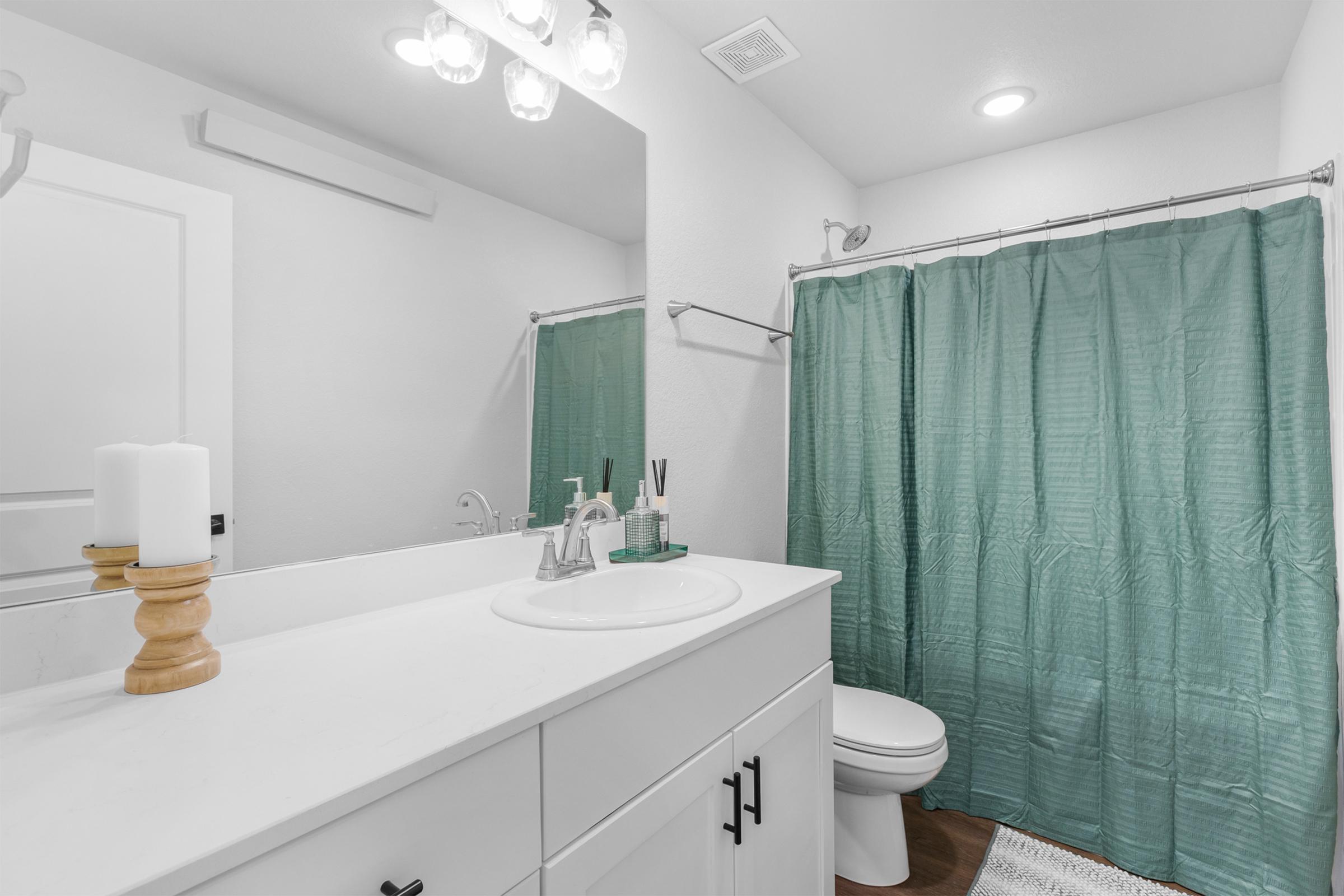 A bright and modern bathroom featuring a white countertop with a sink, a wooden candle holder with candles, a green shower curtain, a toilet, and a large mirror reflecting the space. The walls are light-colored, and the bathroom has a clean, minimalistic design.