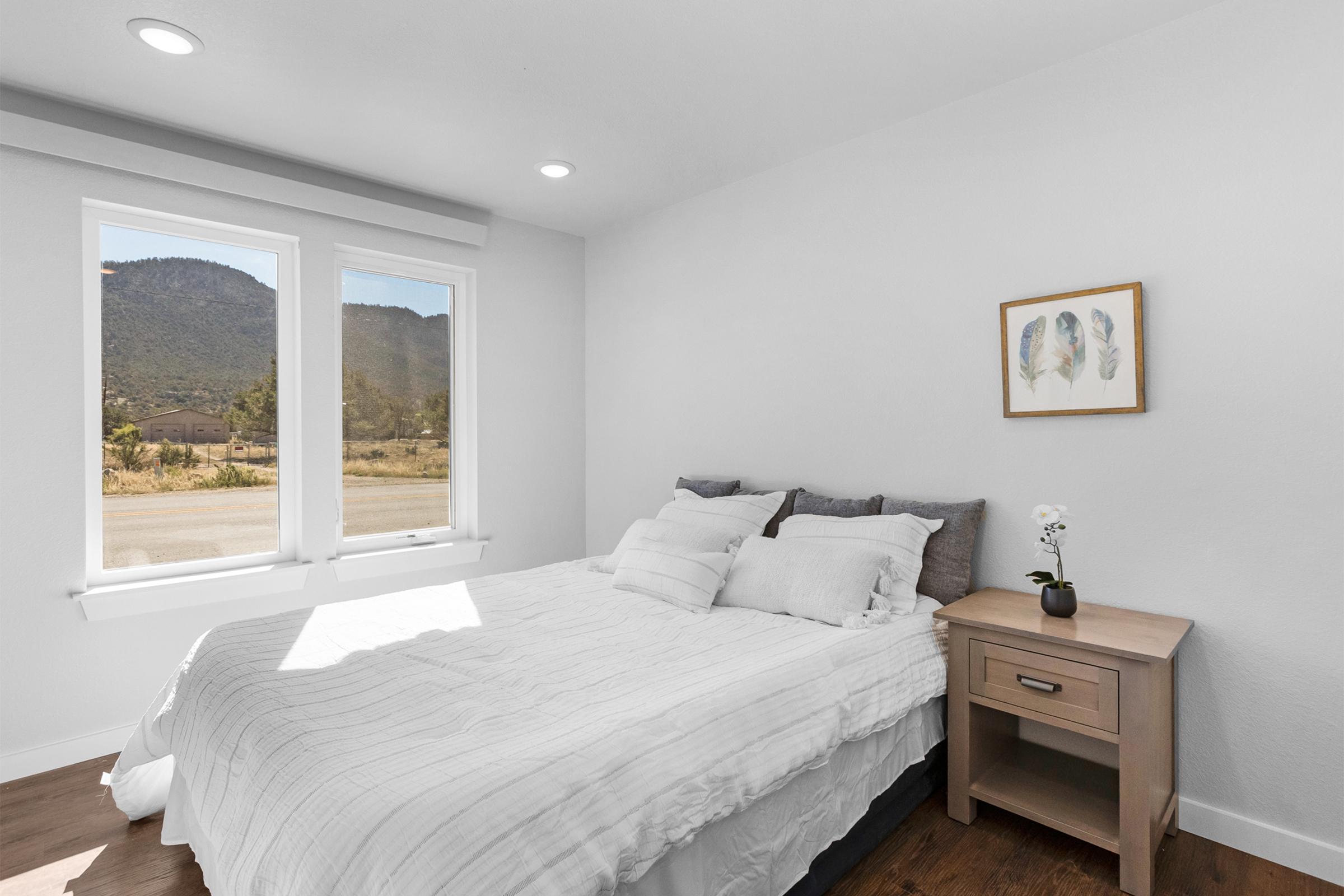 Cozy bedroom featuring a neatly made bed with white bedding and decorative pillows. A small wooden nightstand holds a potted orchid. Two large windows allow natural light to brighten the room, with a scenic mountain view outside. A framed artwork with feathers decorates the wall.
