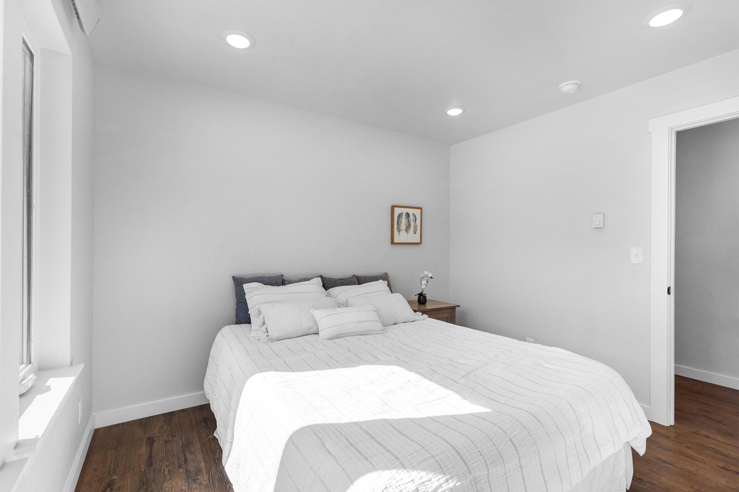 A cozy bedroom featuring a neatly made bed with multiple pillows, a small bedside table with a vase of flowers, and a framed piece of artwork on the wall. The room has light gray walls and warm wood flooring, illuminated by recessed lighting, creating a calm and inviting atmosphere.