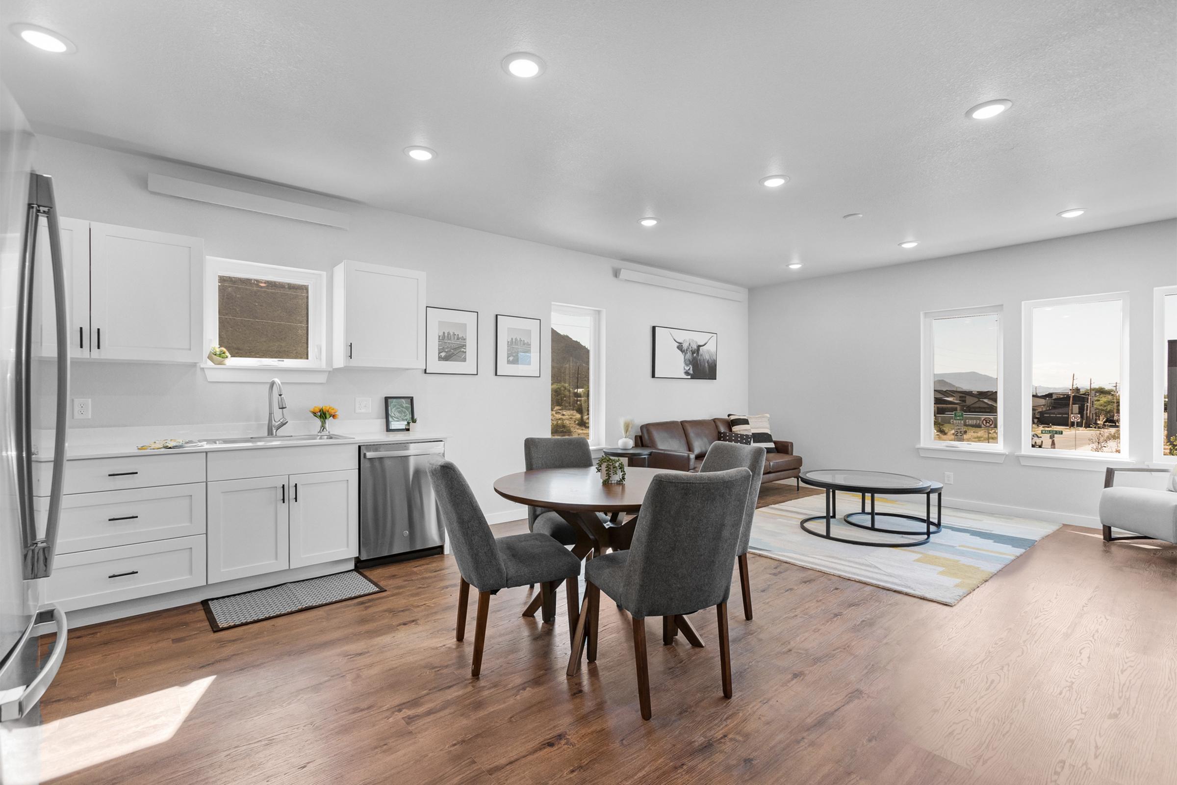 Modern open-concept living space featuring a kitchen with white cabinets, a dining area with a round table and gray chairs, and a cozy living area with a brown couch. Large windows let in natural light, showcasing a view of the outdoors. Minimalist decor with framed pictures on the walls.