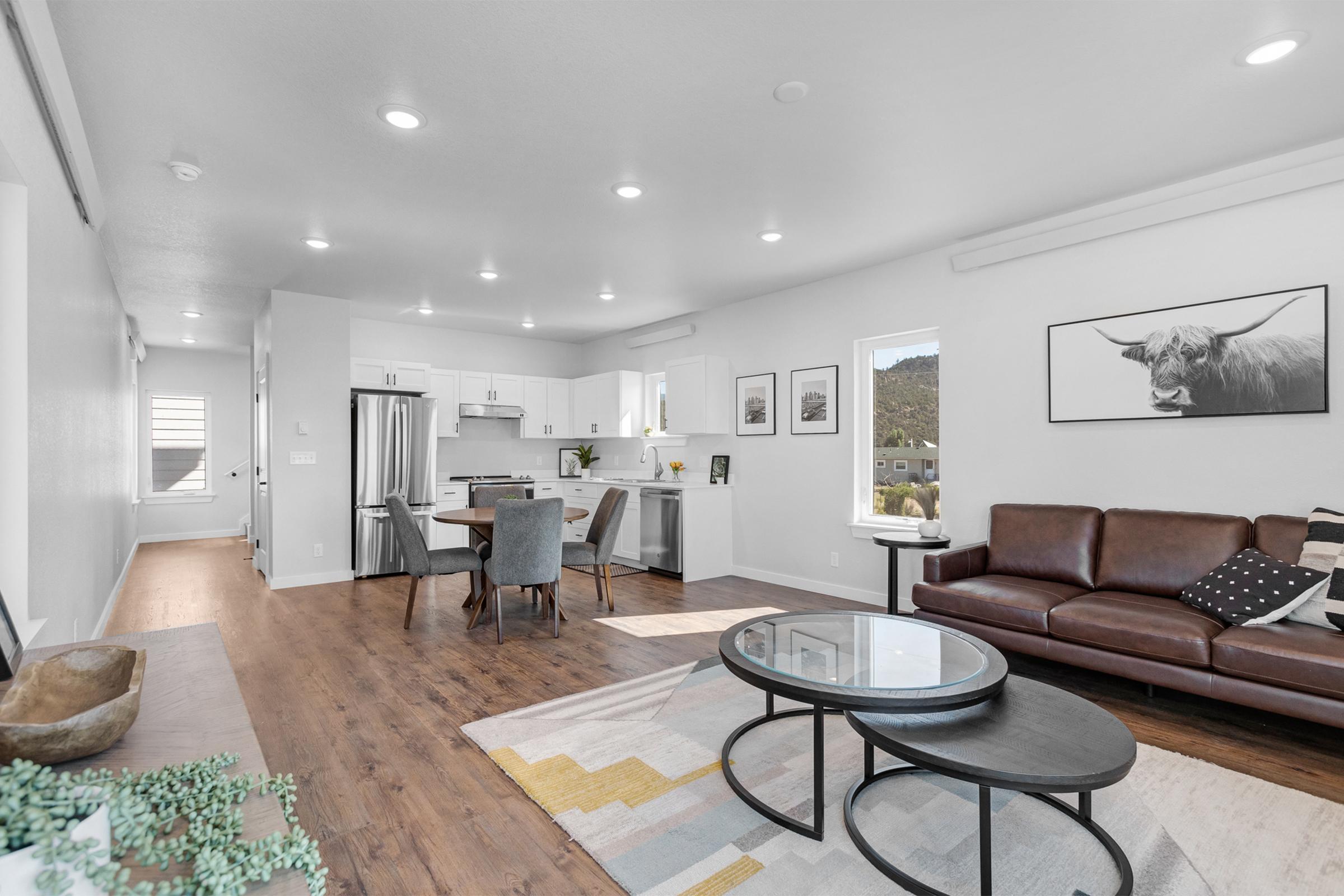 Modern open-concept living space featuring a well-lit kitchen with stainless steel appliances, a dining area with a table and chairs, and a cozy living room with a brown leather sofa. Decorative elements include artwork on the walls, a patterned rug, and a round coffee table.