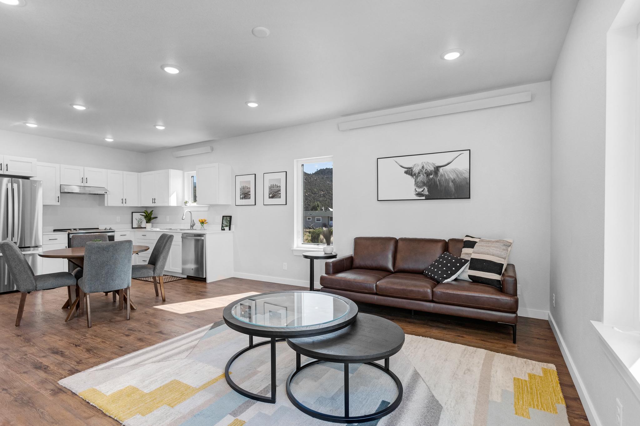 A modern, bright living space featuring a brown leather sofa, circular coffee table, and gray chairs. The open kitchen area includes white cabinetry and stainless steel appliances. Decor includes a wall-mounted artwork of a bull and minimalistic decor elements. Natural light fills the room through large windows.