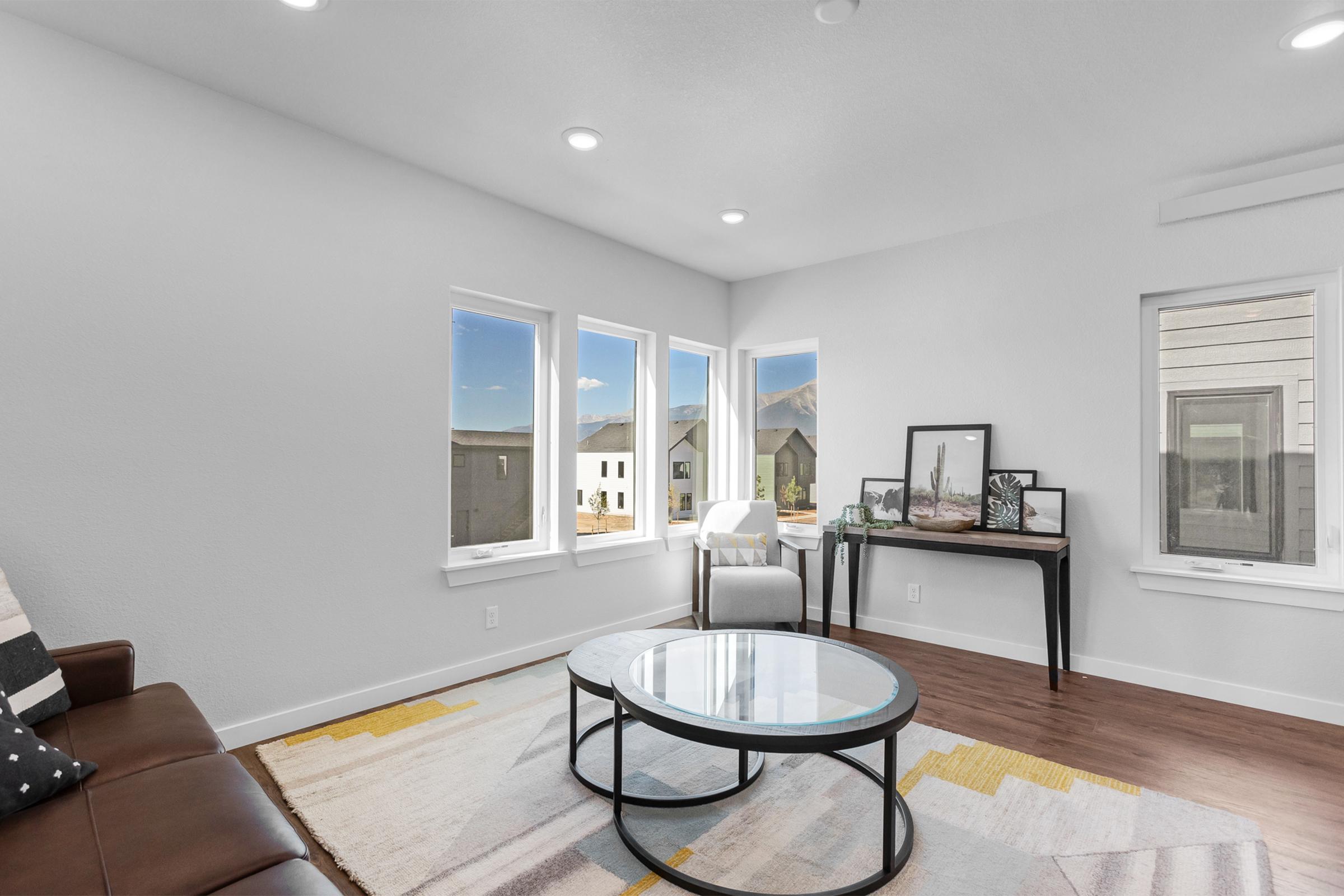 A bright, modern living room featuring a brown leather sofa, a circular glass coffee table, and a small table with framed photos. Large windows provide natural light and a view of nearby houses and mountains in the background. The walls are painted white, and the flooring is hardwood.