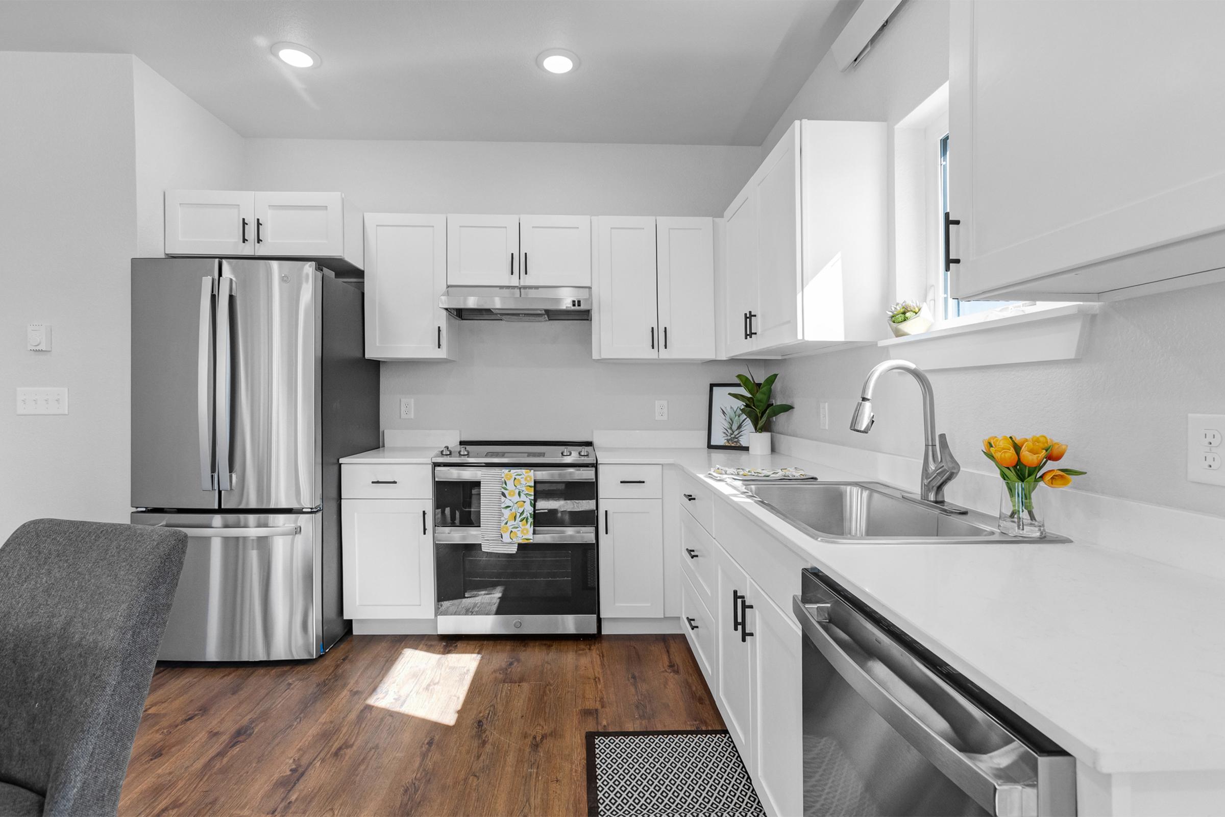 Modern kitchen featuring stainless steel appliances, including a refrigerator and oven. Bright white cabinetry complements the light-colored countertops. A sink with a modern faucet is situated beside a window. A small vase with yellow tulips adds a pop of color, and there's a gray chair in the foreground.