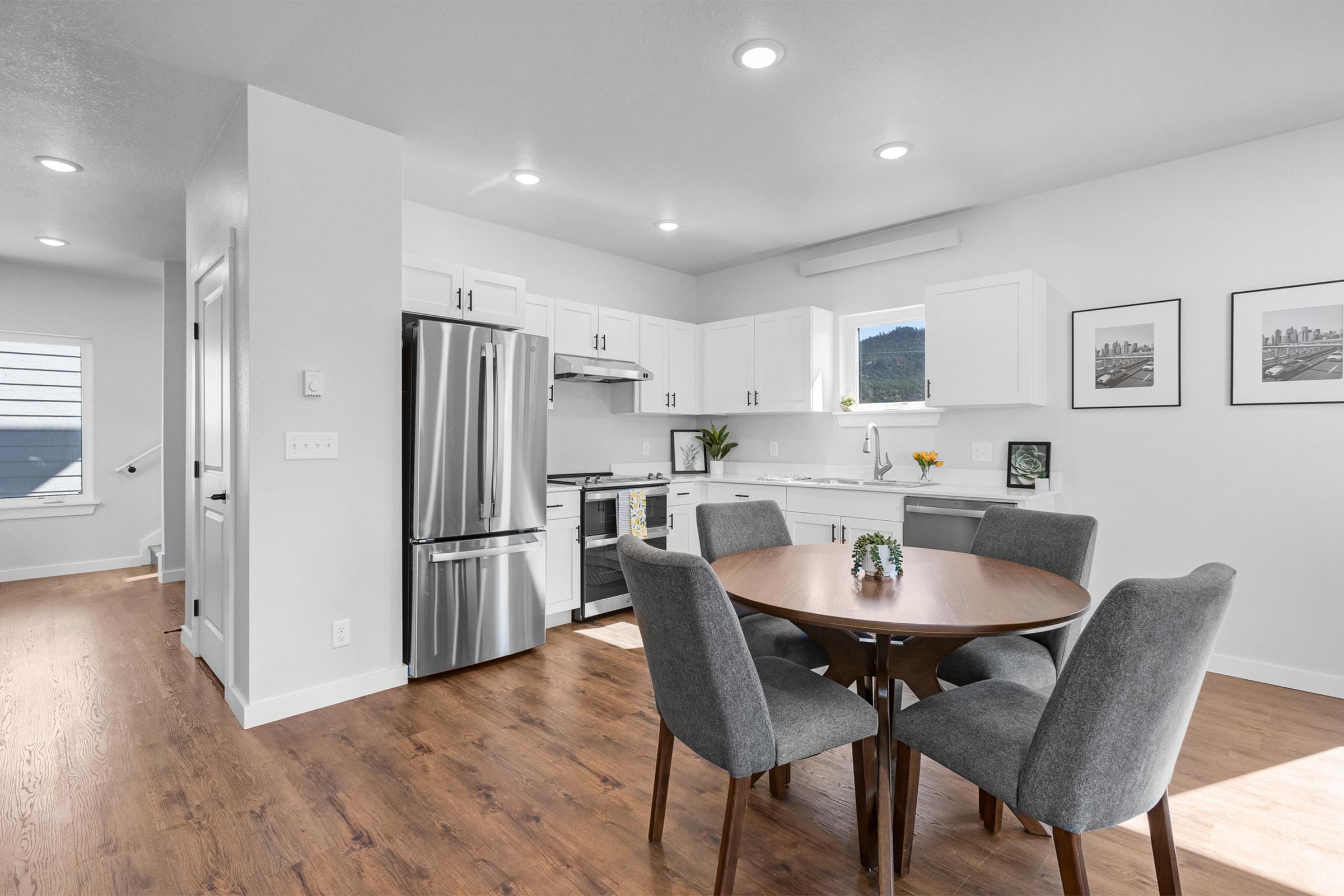 A modern kitchen and dining area featuring a round wooden table with gray upholstered chairs, stainless steel appliances, and white cabinetry. The room is bright with natural light, showcasing hardwood flooring and minimalist decor, including framed pictures on the wall.