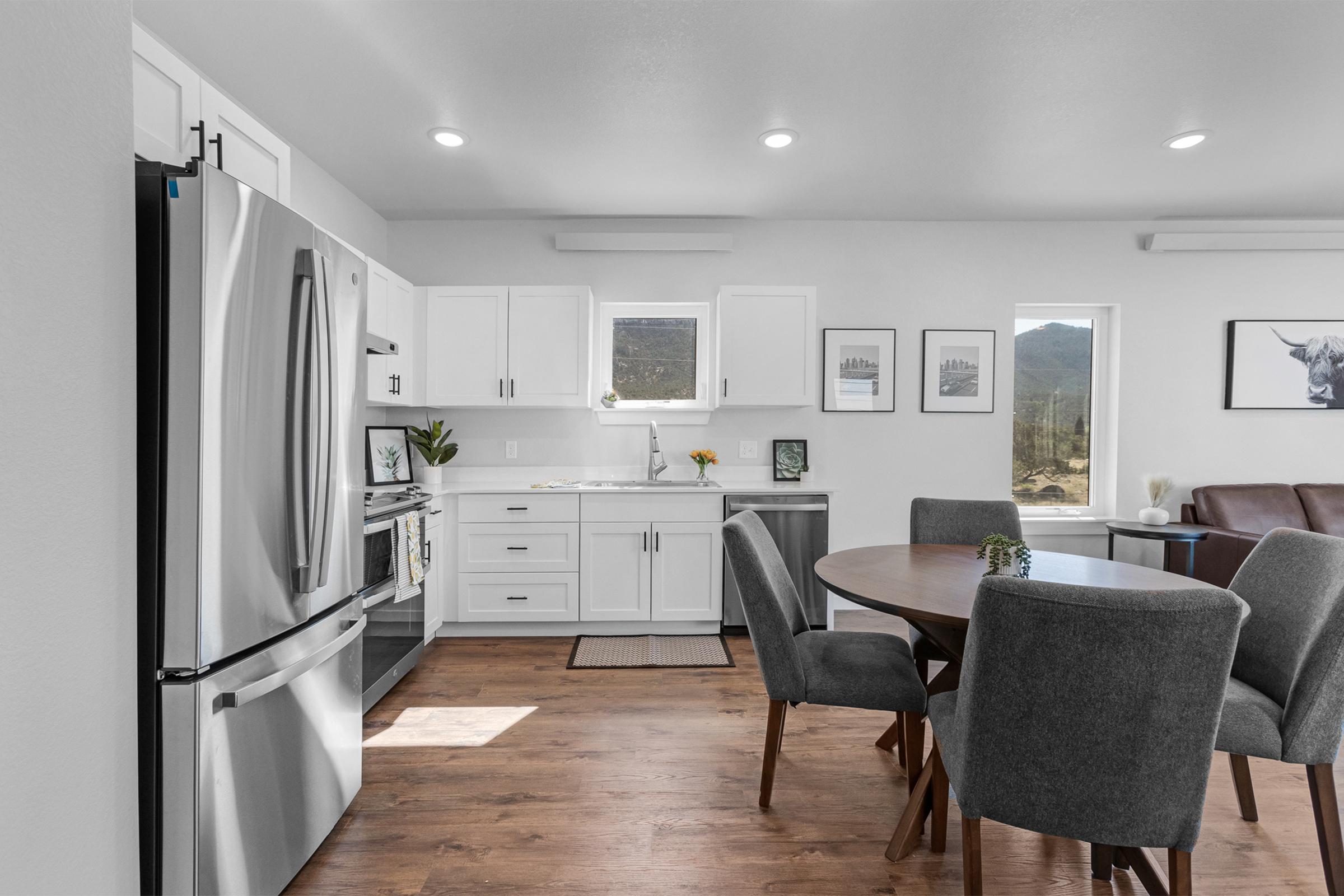 Bright modern kitchen with stainless steel appliances, including a refrigerator and stove. White cabinetry complements a round dining table surrounded by gray upholstered chairs. Natural light streams through windows, offering a view of a mountainous landscape in the background.
