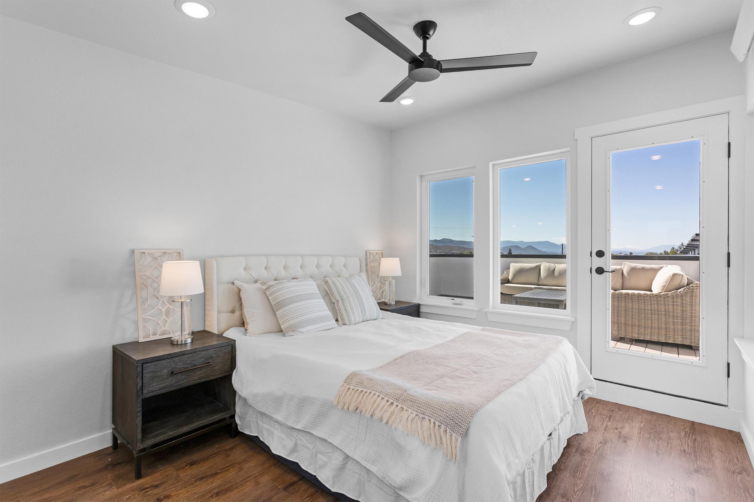 A modern bedroom featuring a queen-sized bed with white bedding, two bedside lamps, and a wooden nightstand. Large windows provide a view of mountains and a balcony, with a cozy seating area visible outside. The room has a light and airy atmosphere, complemented by a ceiling fan and neutral decor.