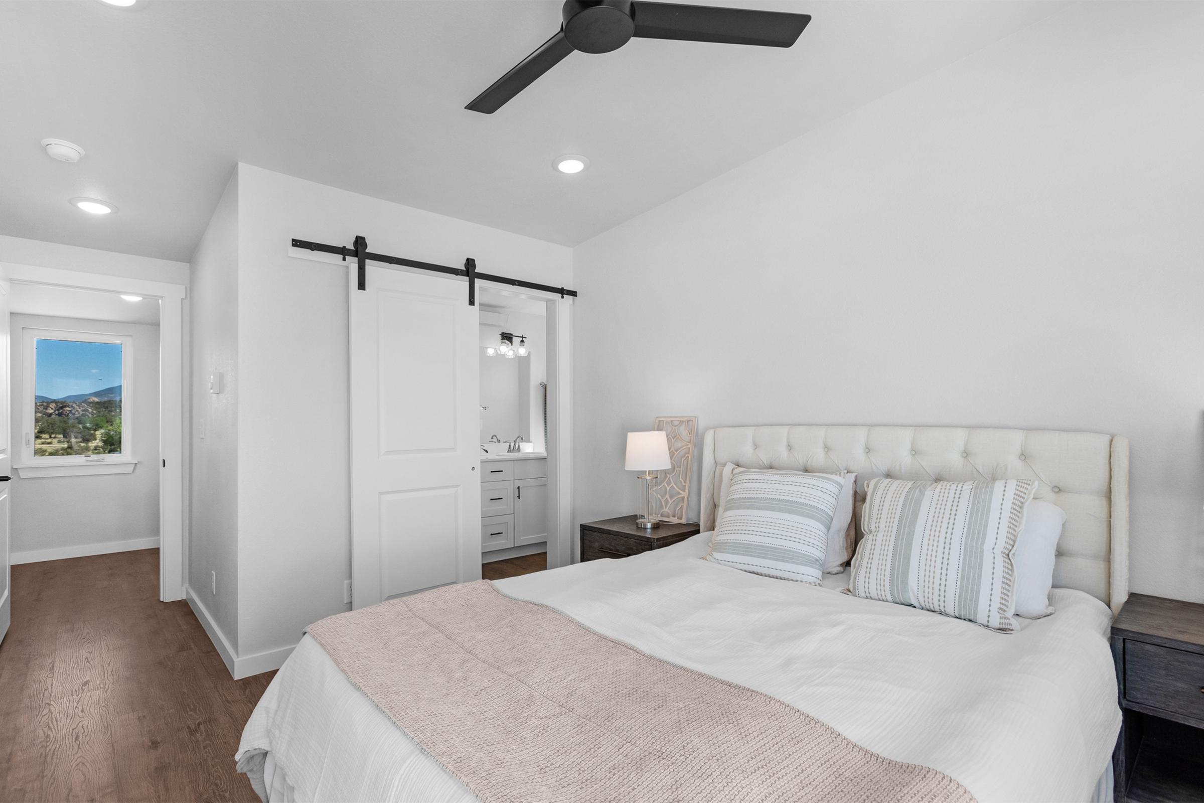 A cozy, modern bedroom featuring a light-colored bed with decorative pillows, a nightstand with a lamp, and a sliding barn door leading to a bathroom. Natural light filters in from a window, creating a warm and inviting atmosphere. The walls are painted white, and there is a fan on the ceiling.