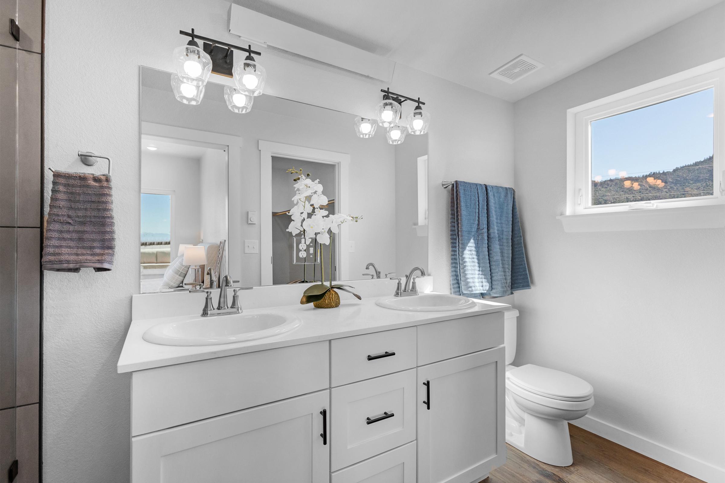 A modern bathroom featuring a double vanity with two sinks, elegant lighting fixtures above, and a decorative vase with an orchid. The walls are light-colored, and there is a window showing an outdoor view. Blue towels hang on the wall, and a toilet is visible in the corner.