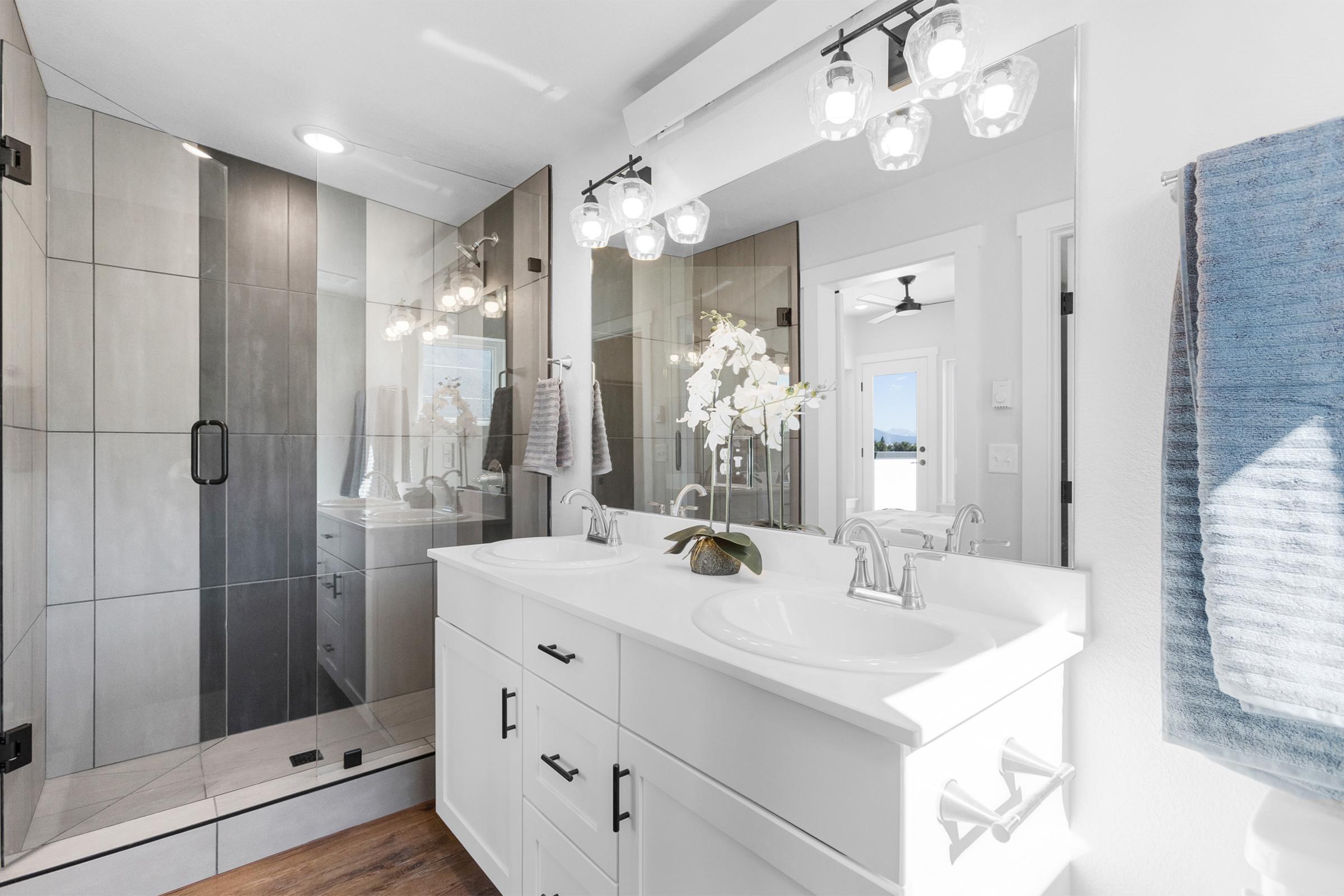 Modern bathroom featuring a double sink vanity with white cabinetry, a large mirror, and stylish light fixtures. A glass-enclosed shower with gray tile is visible, along with a blue towel hanging on the wall and an orchid plant on the countertop. The overall decor is sleek and contemporary.