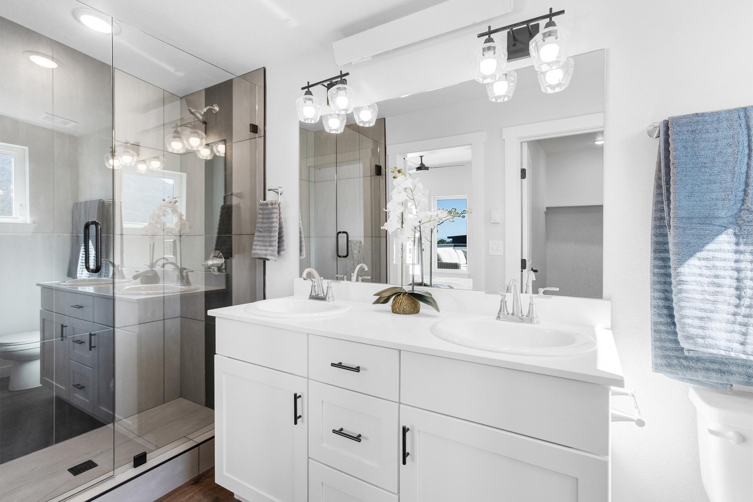 A modern bathroom featuring a double vanity with two sinks, elegant light fixtures, and a glass shower enclosure. The walls are painted white, and there are decorative elements like a vase with an orchid. A towel hangs on a rack, and a glimpse of a bedroom is visible through a doorway.