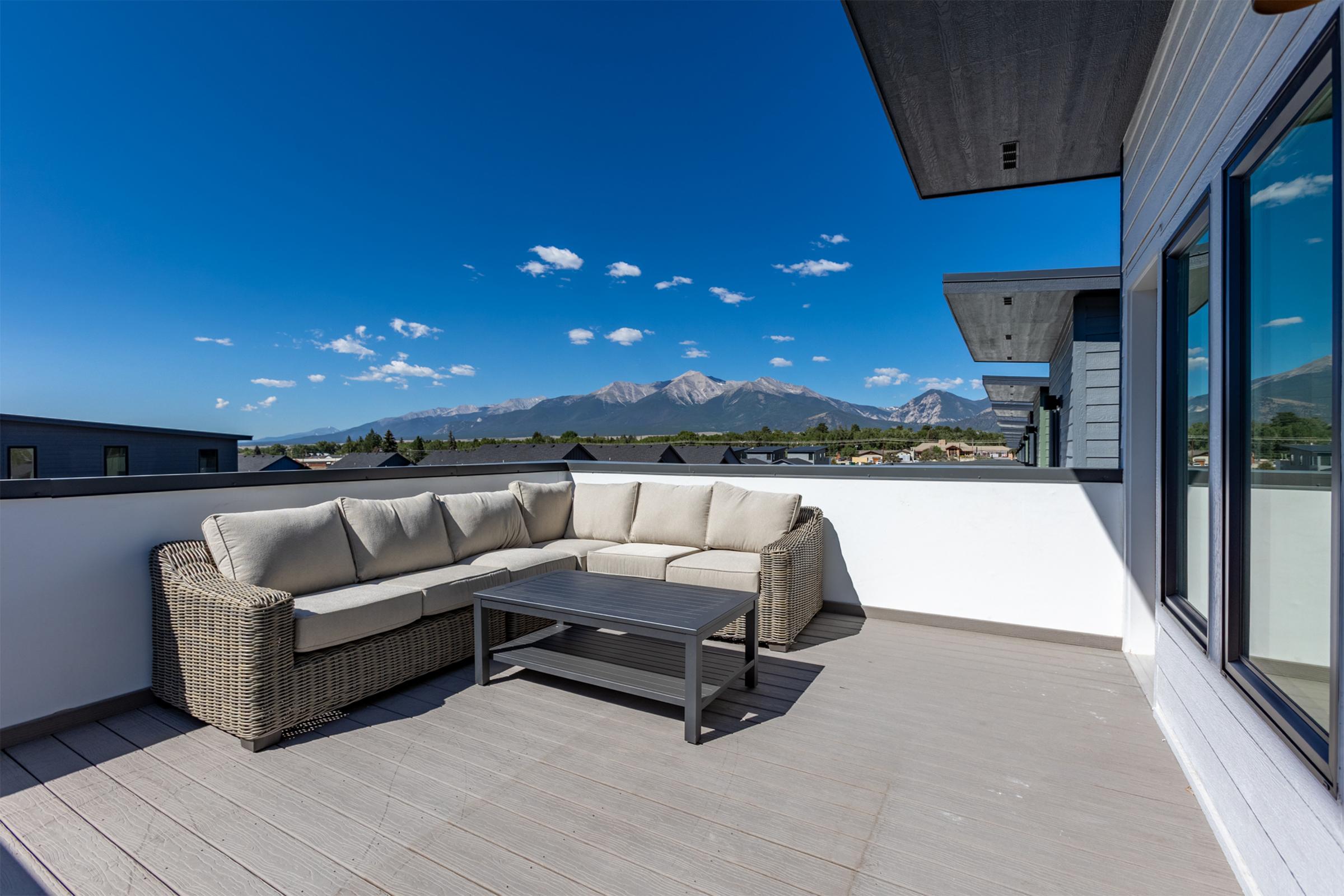 A spacious rooftop terrace with a comfortable sectional sofa and a coffee table, featuring a stunning view of distant mountains under a clear blue sky. The area is designed for relaxation and enjoyment of the scenic landscape.