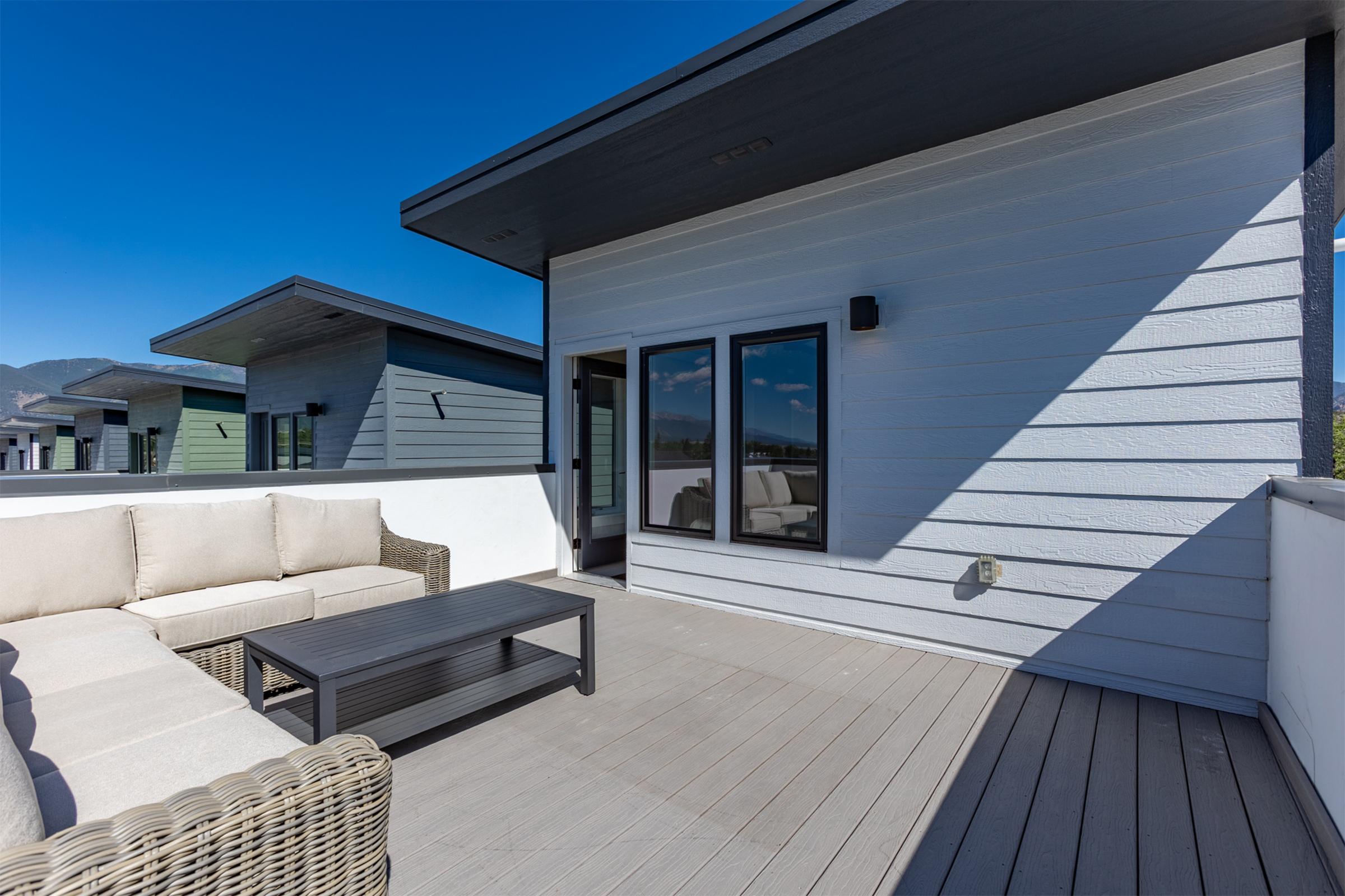 A modern rooftop terrace featuring a light-colored sectional sofa and a dark coffee table. The area is surrounded by stylish walls and has large windows, with a clear blue sky visible above. The design reflects a contemporary aesthetic with clean lines and a minimalist approach.