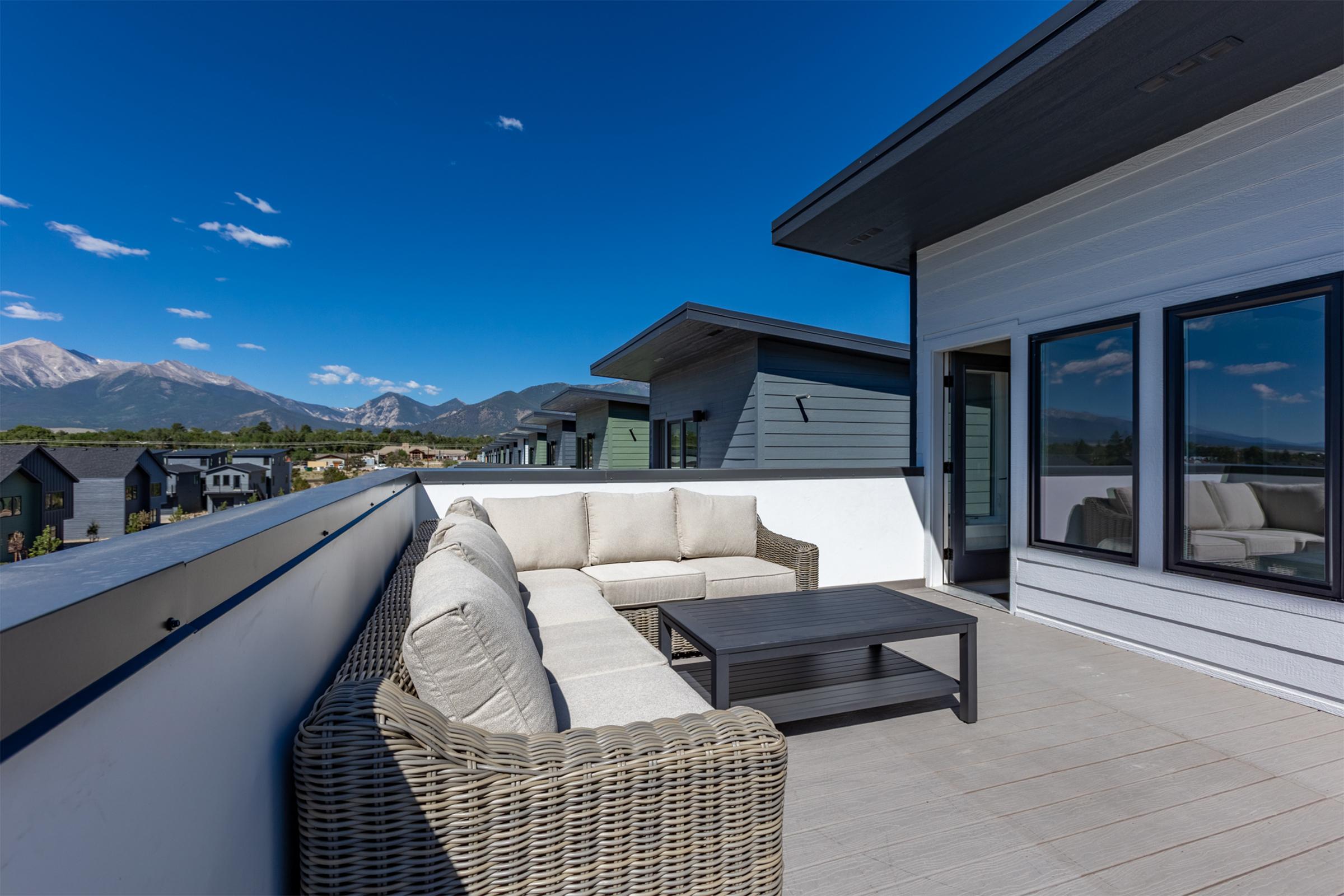 A rooftop terrace featuring a light-colored sectional sofa and a black coffee table. The space is surrounded by modern buildings and offers a view of mountains under a clear blue sky. Bright sunlight enhances the inviting atmosphere of the outdoor seating area.