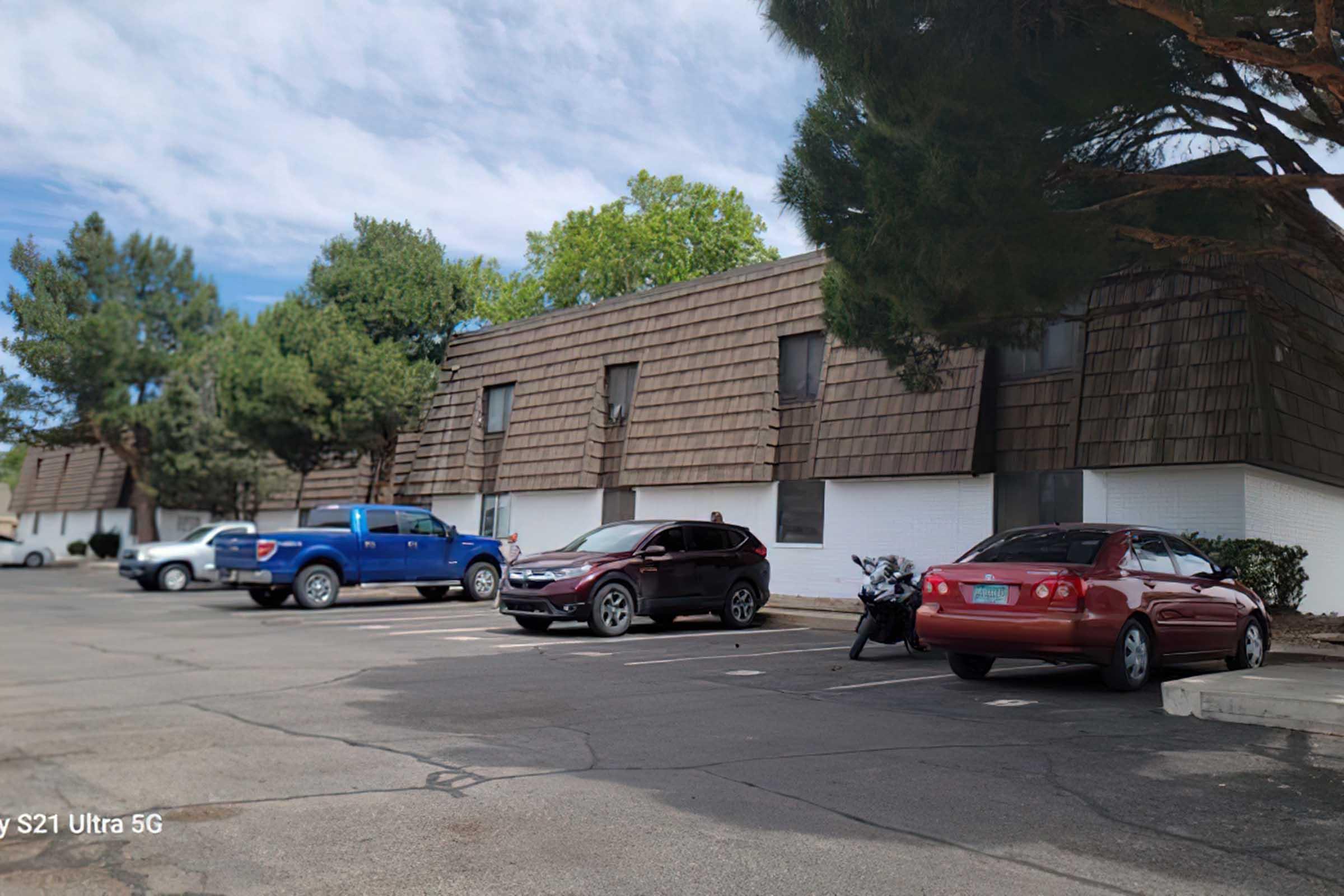 A parking lot with several cars, including a blue truck, a gray SUV, a red car, and a motorcycle. Trees line the area, and there are two buildings with brown shingled roofs in the background. The scene is sunny with a clear sky.