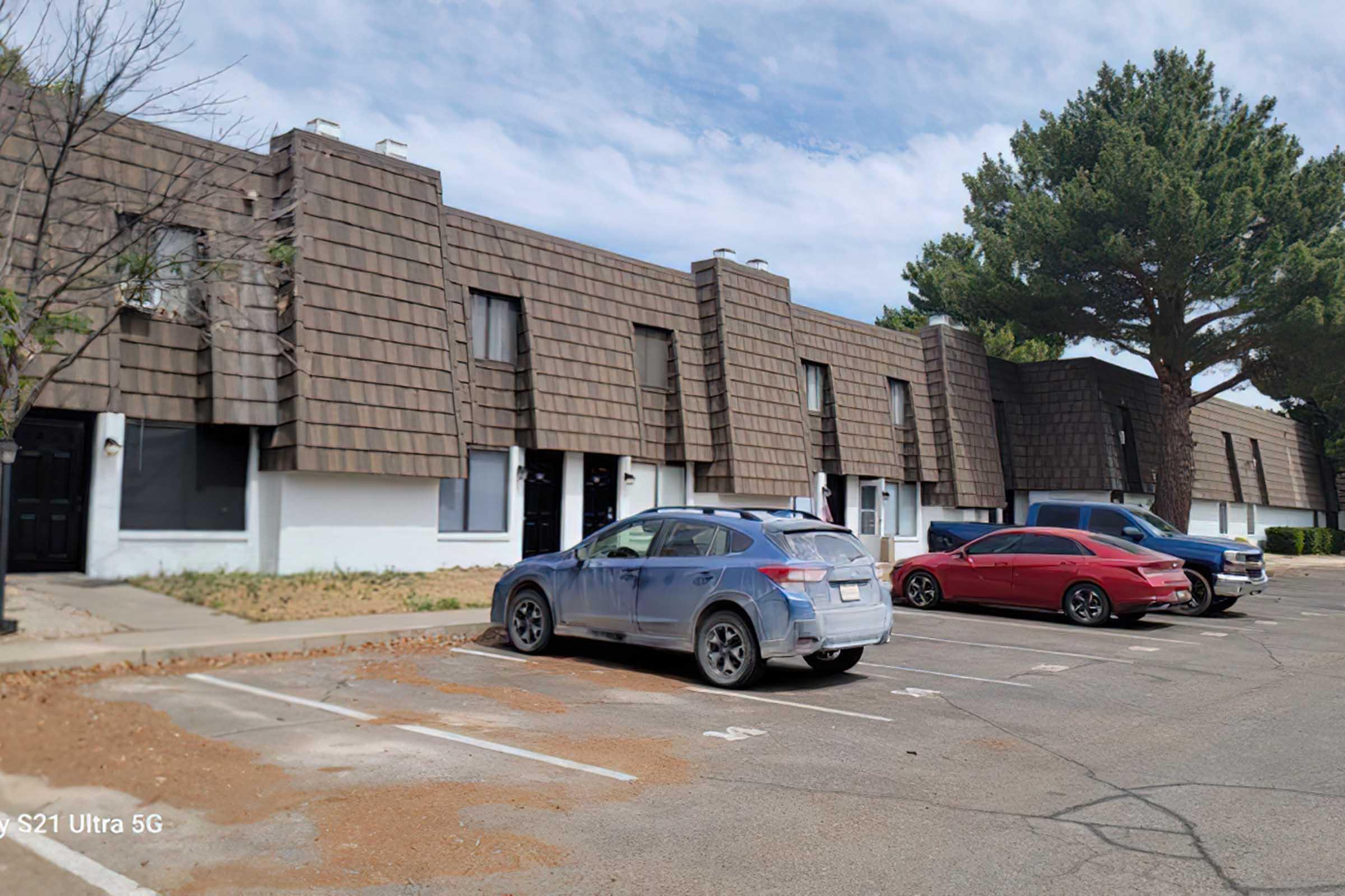 Two-story apartment buildings with a distinctive brown shingle roof and white walls. Several parked cars, including a white SUV and a red sedan, are visible in the foreground. The surrounding area features some trees and a gravelly patch of ground. The sky is clear and the scene is well-lit.