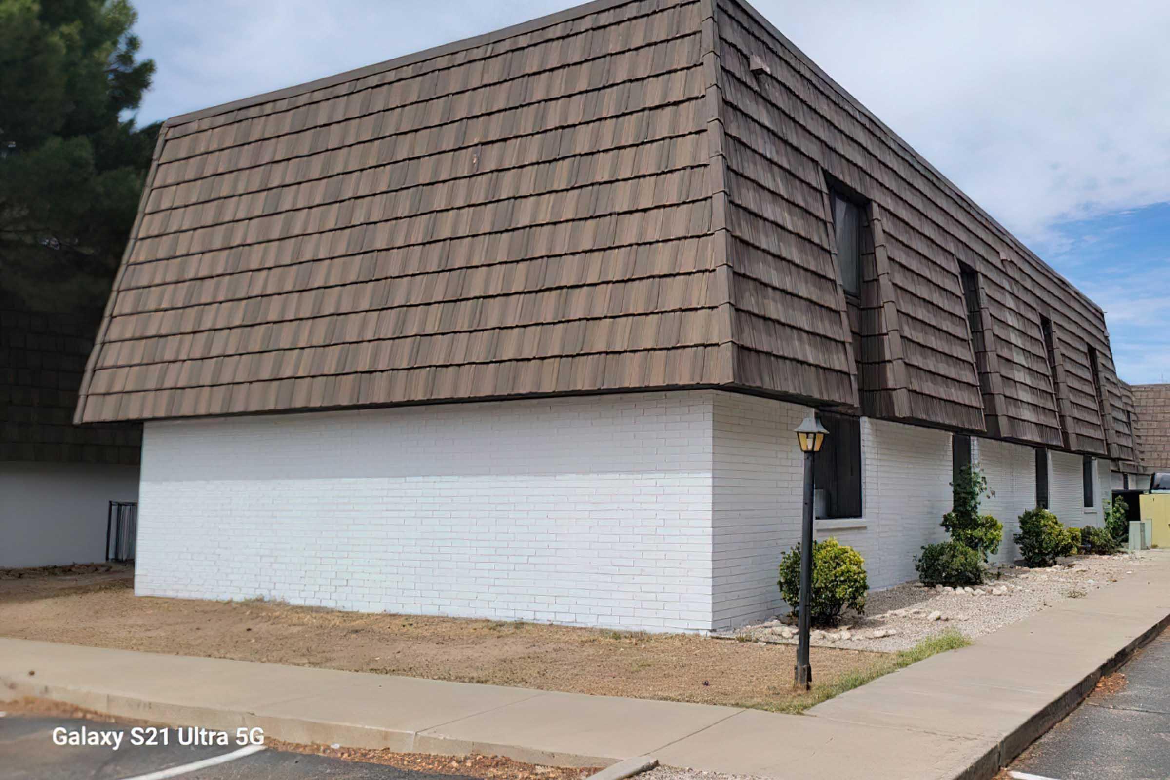 A two-story building featuring a distinctive brown shingled roof and white brick walls. The surrounding area has gravel and small shrubs, with a sidewalk leading up to the entrance. The sky is partly cloudy, suggesting a clear day. A lamppost is visible near the sidewalk.